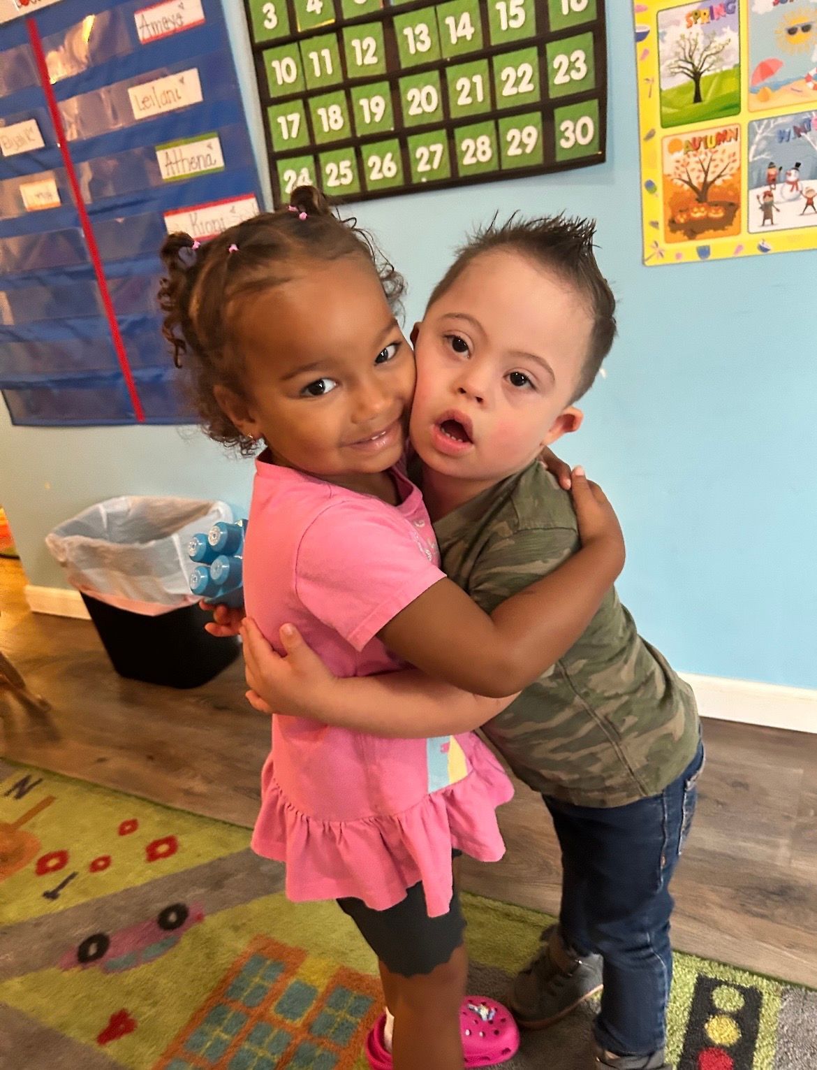 Two children hugging in a colorful classroom, standing on a patterned rug.