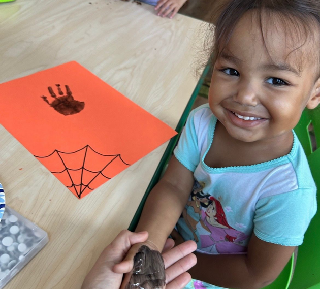 Smiling child at a table with an orange spider craft and handprint, holding a small frog-like toy.