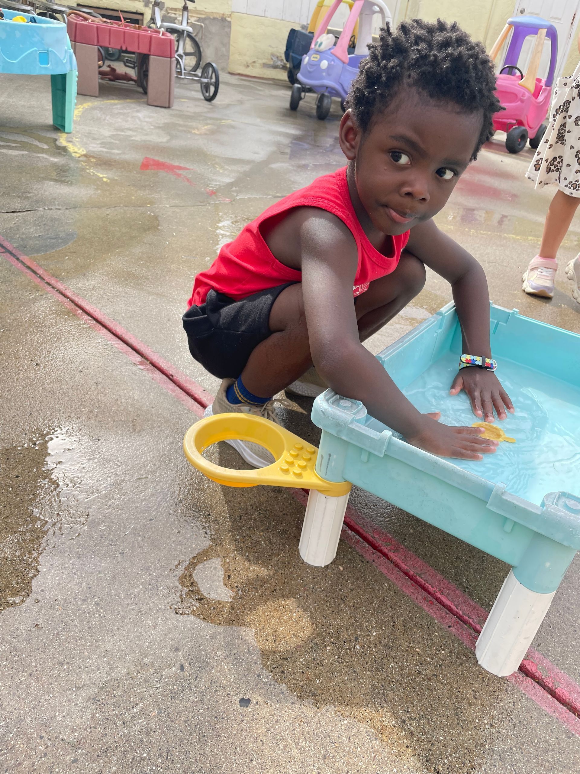A young child in a red tank top and black shorts plays in a blue plastic water table on a concrete outdoor playground.