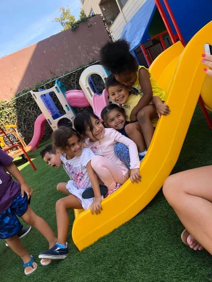 Teacher and four children playing a game at a table in a brightly decorated classroom.