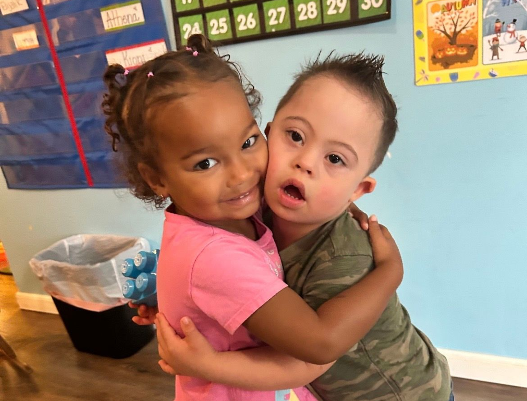 Two children hugging in a classroom, one in pink and one in camouflage, smiling at the camera.