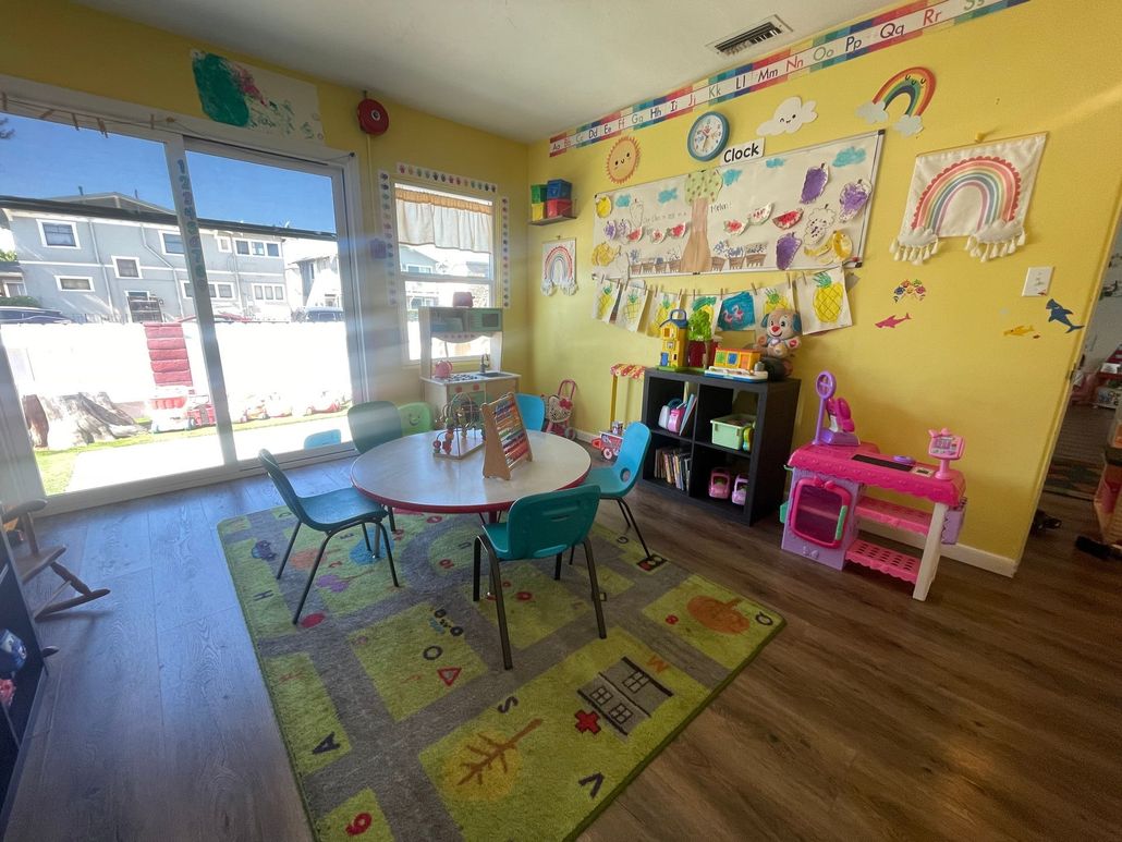 A brightly colored, yellow-walled preschool classroom with a circular table, chairs on a rug, and a toy play kitchen.