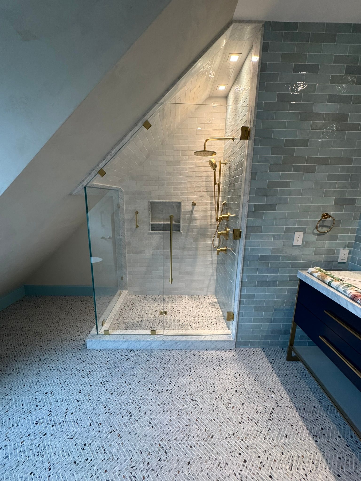 Modern bathroom with angled shower and mosaic tile floor. Gray brick wall and gold fixtures.