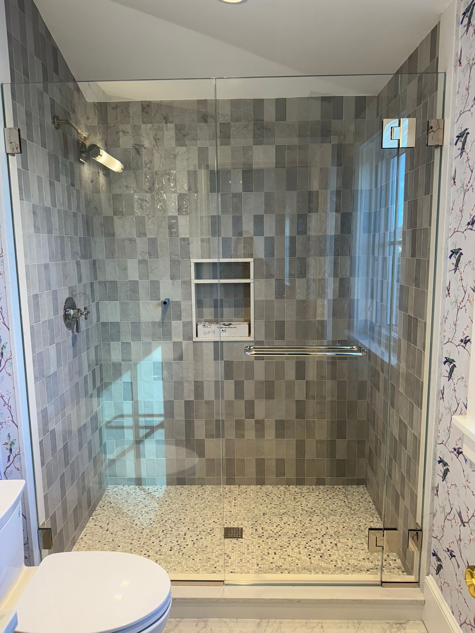A modern shower with gray tile walls, pebble floor, and glass doors. Features a recessed shelf and a grab bar.