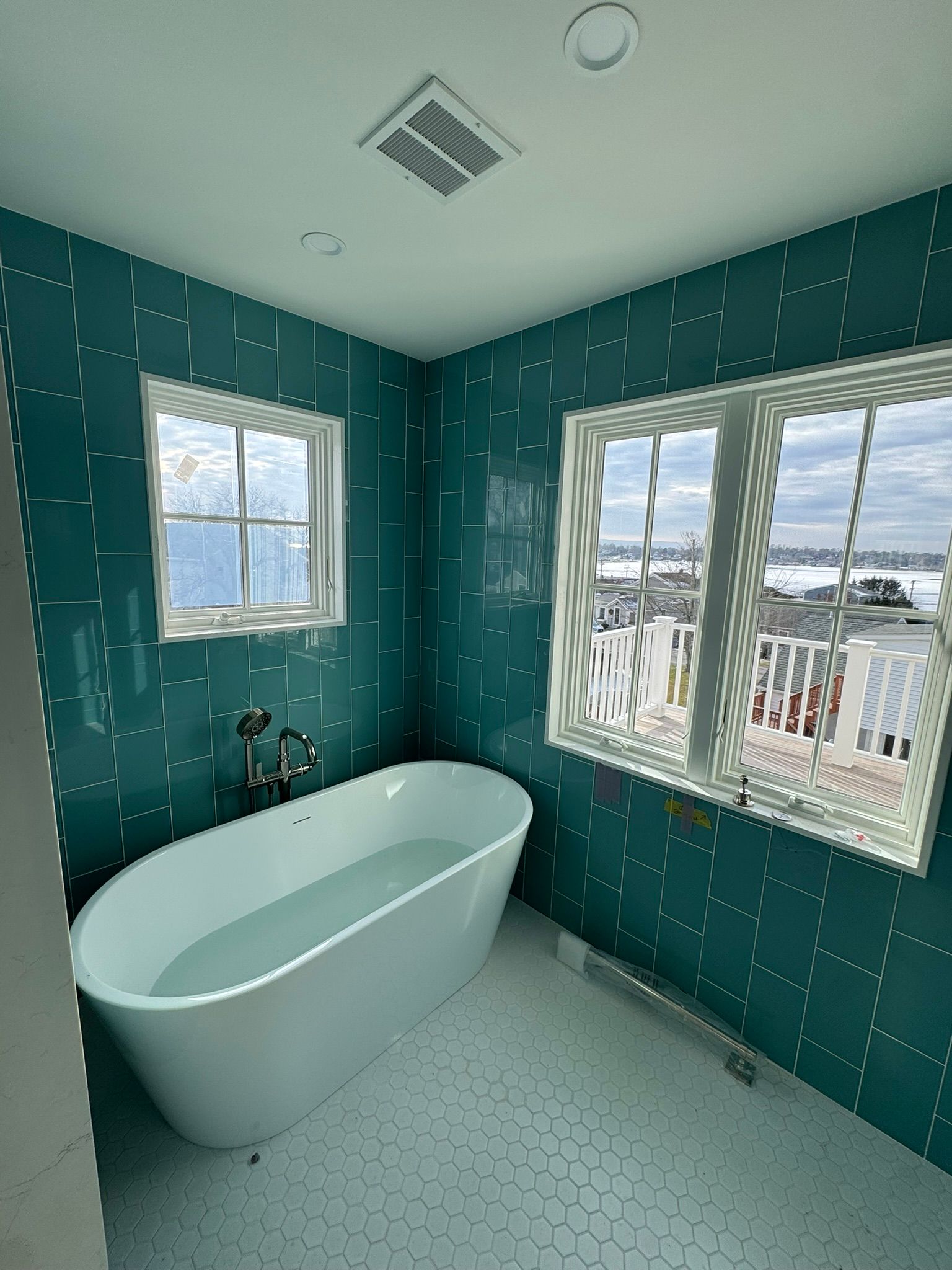 Teal tiled bathroom with a white soaking tub by two windows, natural light.