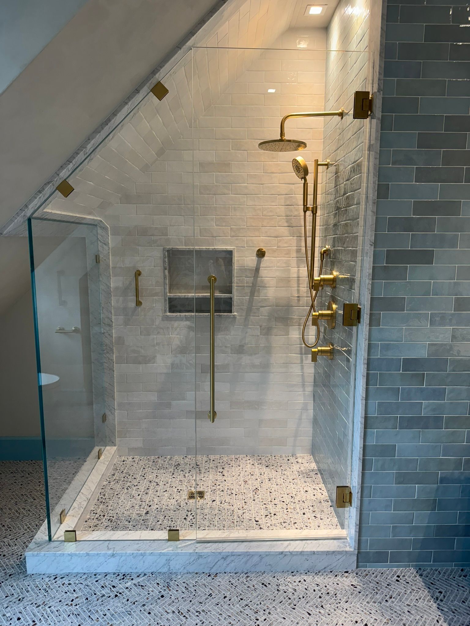 Modern shower with glass doors, gold fixtures, and light gray brick tile. Pebble floor, recessed niche.