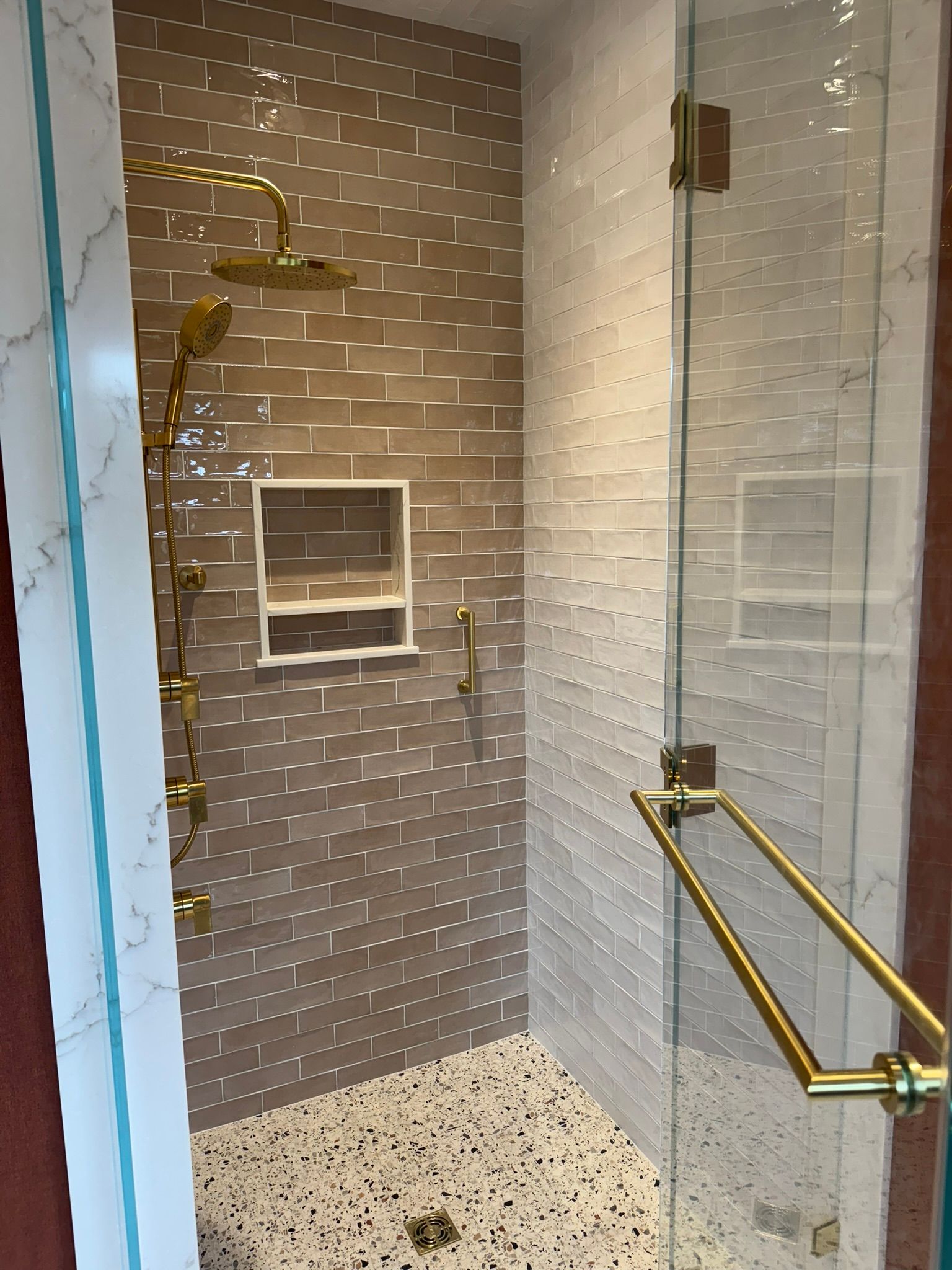 Bathroom with gold fixtures, glass shower door, and two-toned brick tile walls; pebble floor.