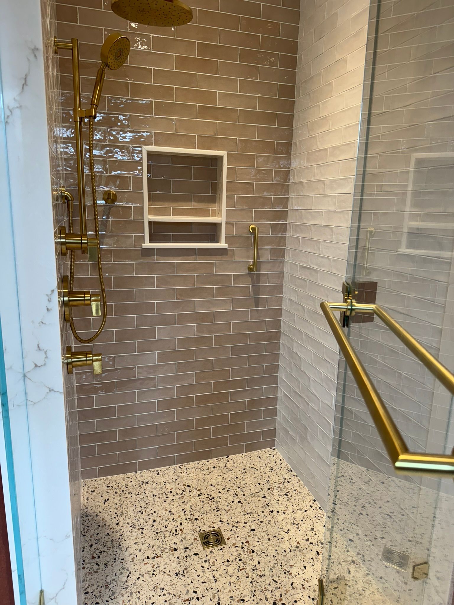Shower with gold fixtures, brown tiled walls, and terrazzo floor.