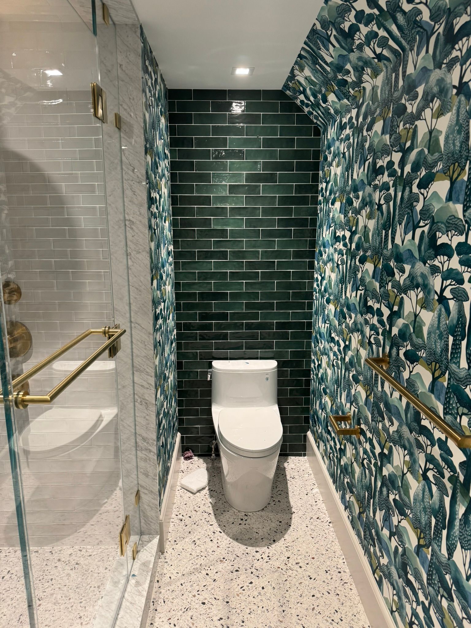 Small bathroom with toilet and shower, green tile and patterned wallpaper, gold accents, terrazzo floor.