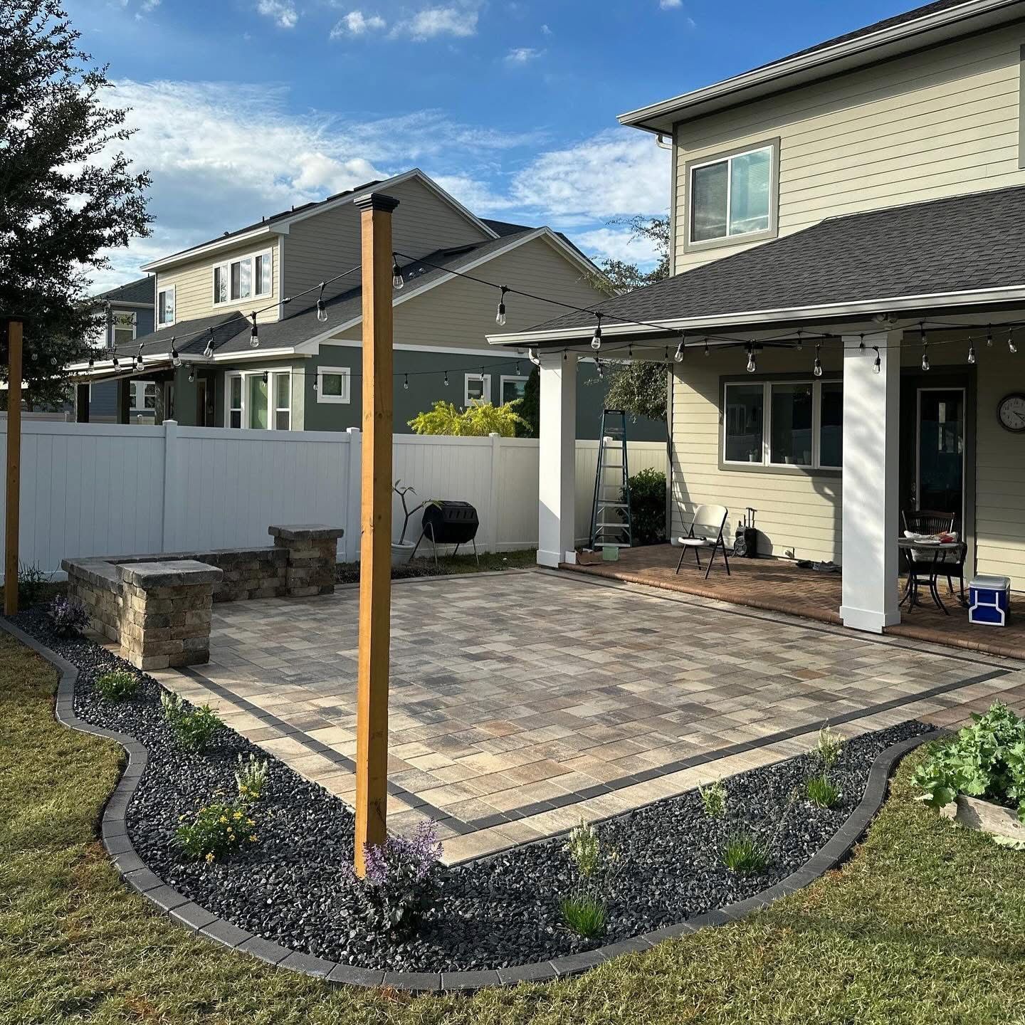 Backyard patio with stone pavers, white fence, and two wooden posts beside a beige house