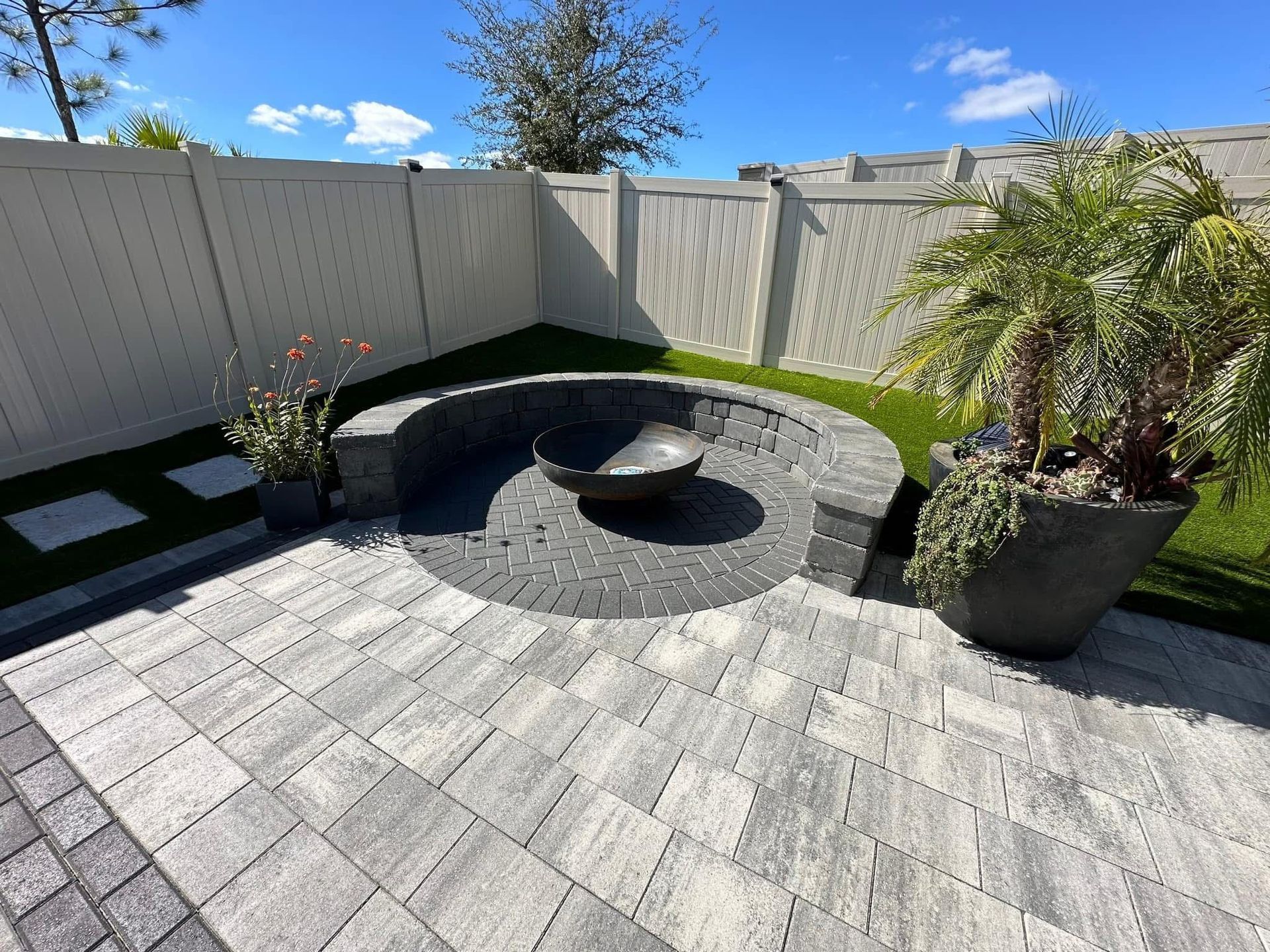 Modern patio with gray pavers, curved black bench, fire pit, and potted plants by a white fence under blue sky