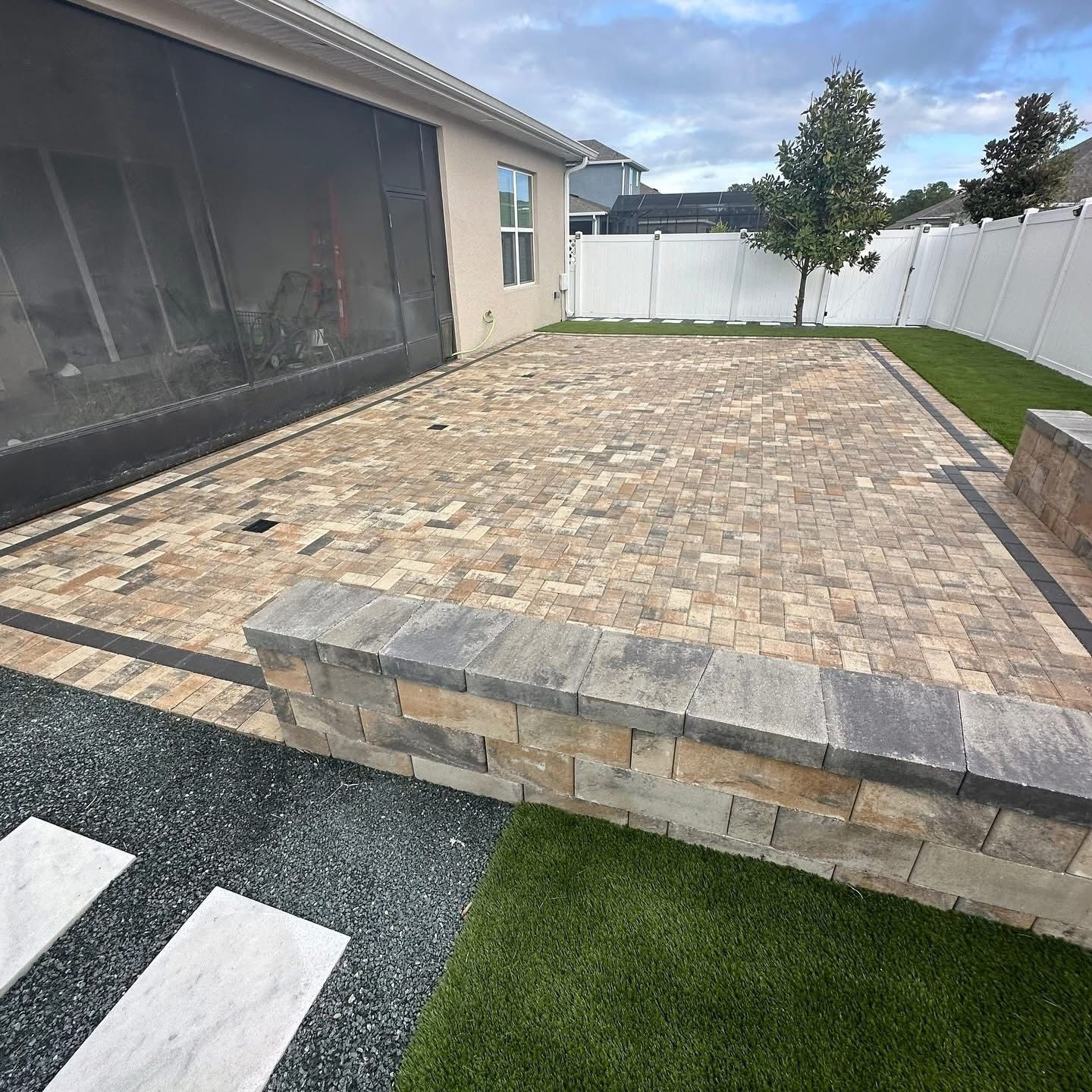 Raised stone patio in a backyard with white fence, grass border, and house siding.