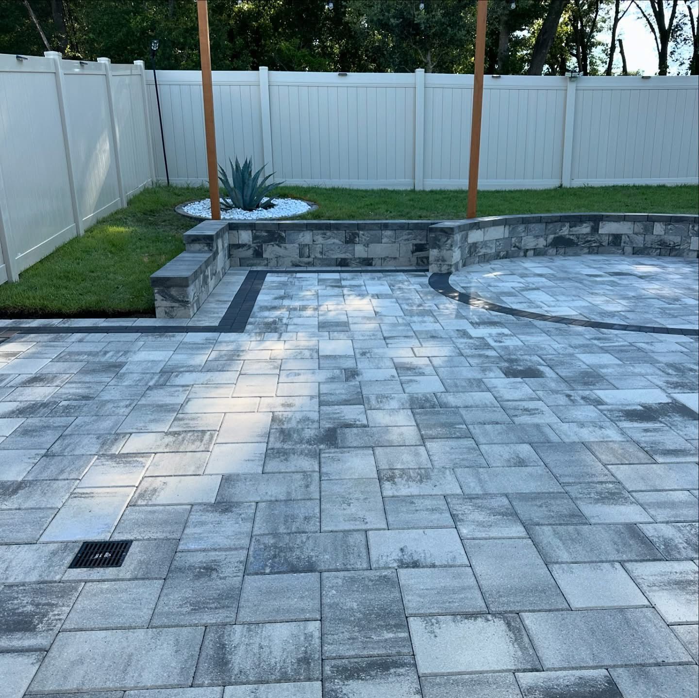Stone patio and retaining wall in a fenced backyard with grass and trees.