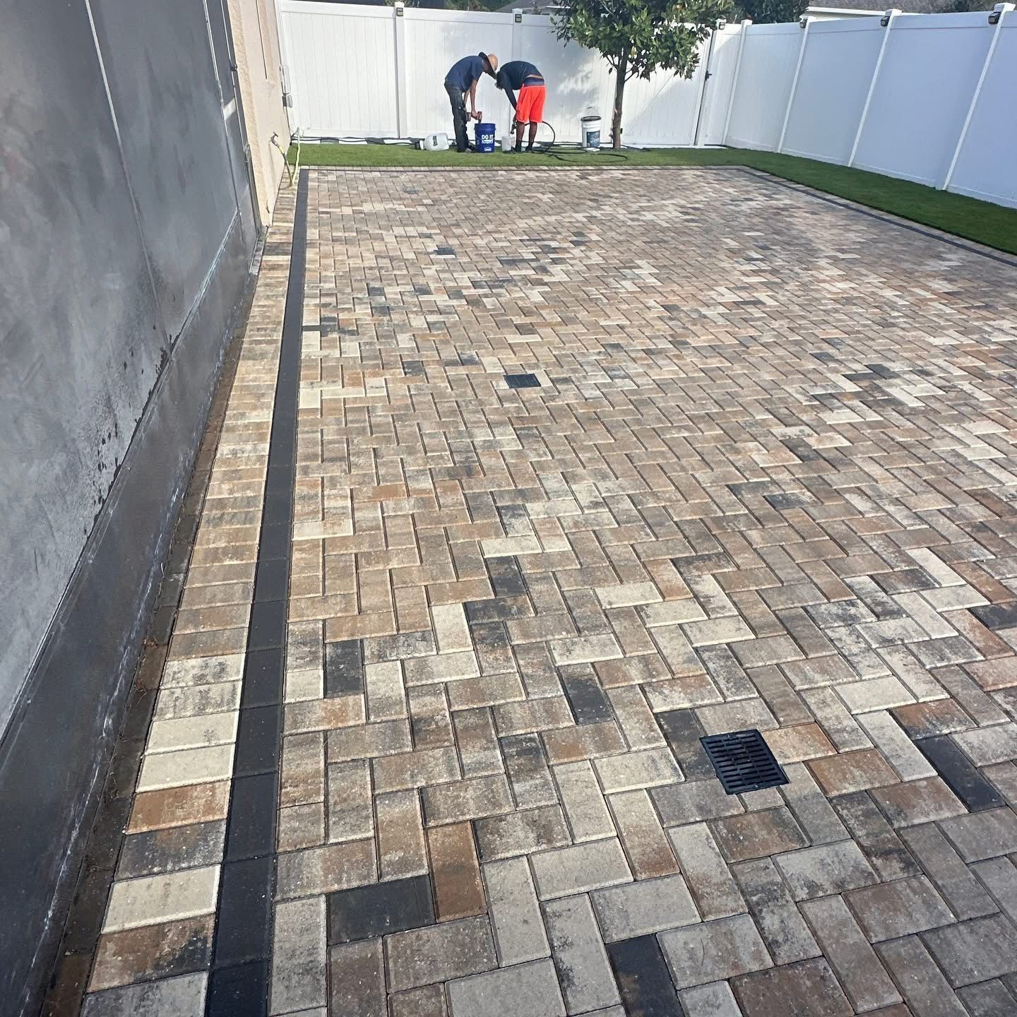 Paved backyard patio with a white fence and two people working near the far end.