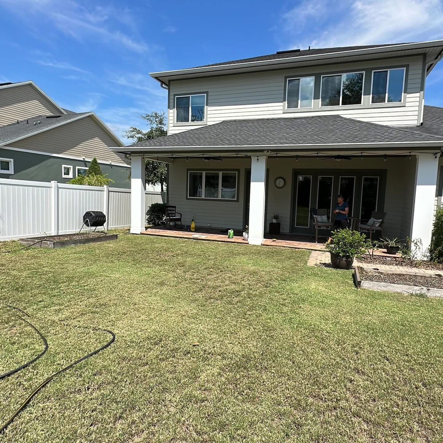 Backyard view of a two-story house with a covered patio, green lawn, and white fence under a blue sky