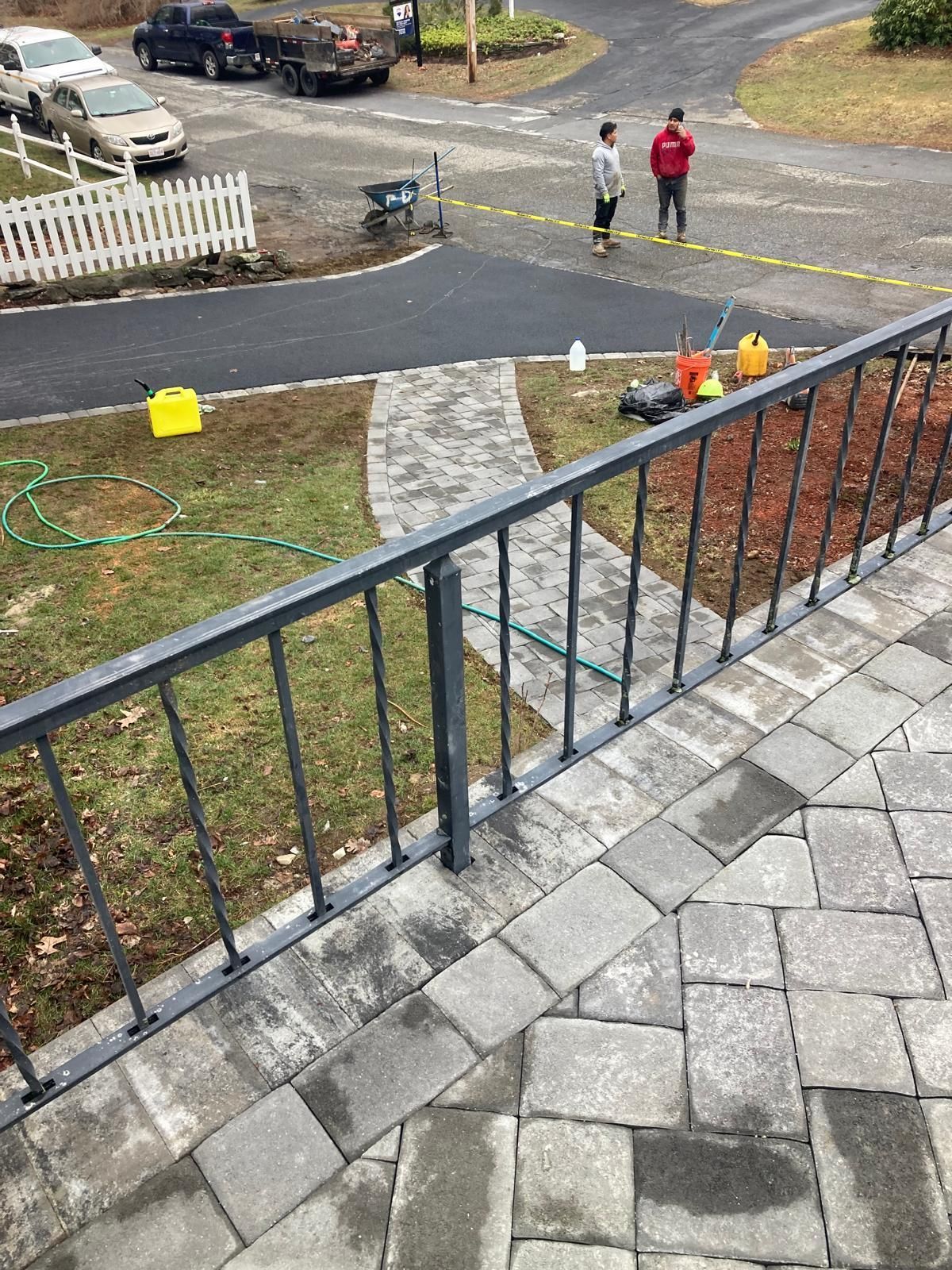 Stone walkway and metal railing overlooking a driveway, with two people standing near the curb and orange cones.