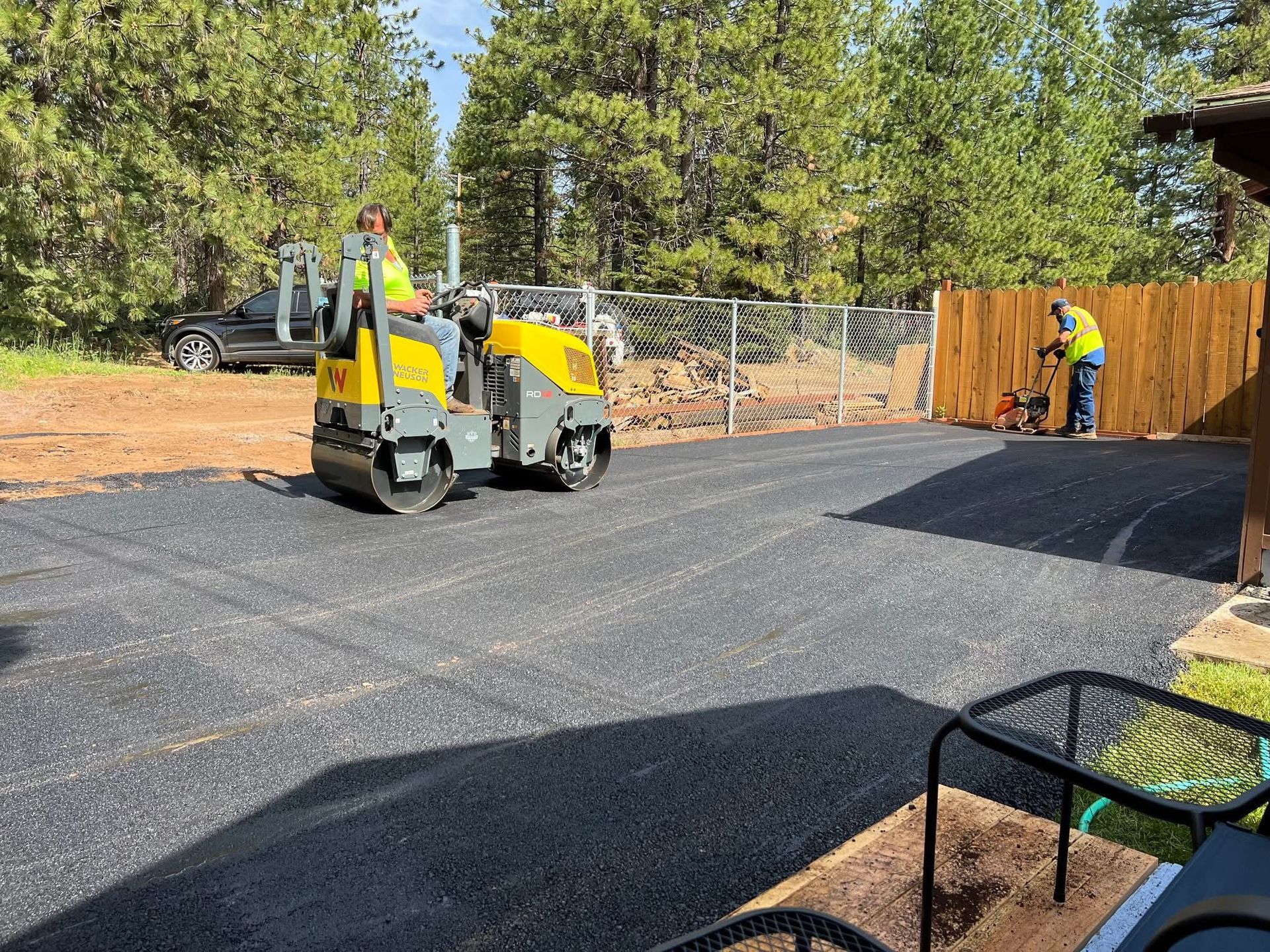 Two workers use yellow road rollers to compact fresh asphalt on a driveway beside a fence and trees.