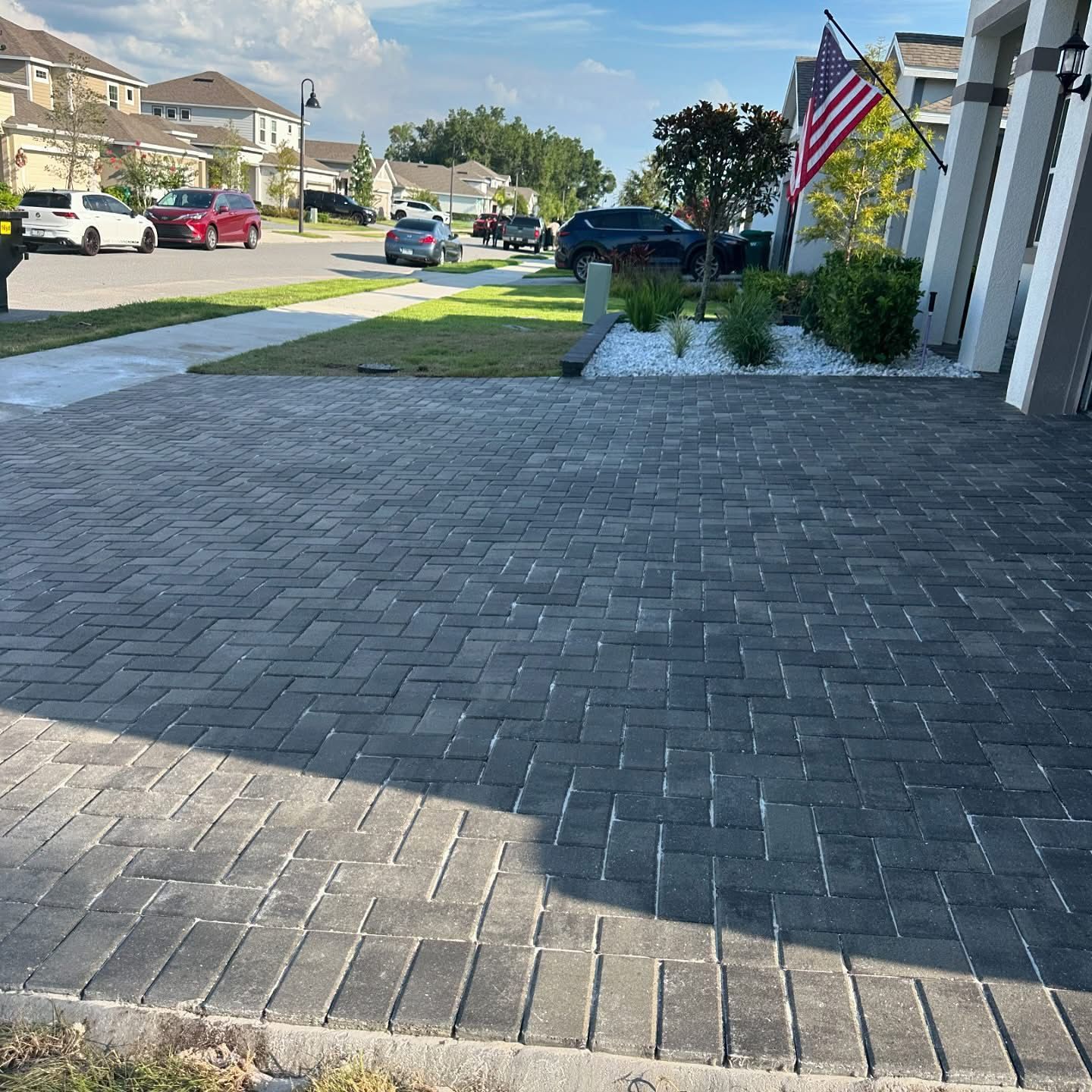 Suburban driveway with brick pavers, parked cars, and an American flag by a row of houses.