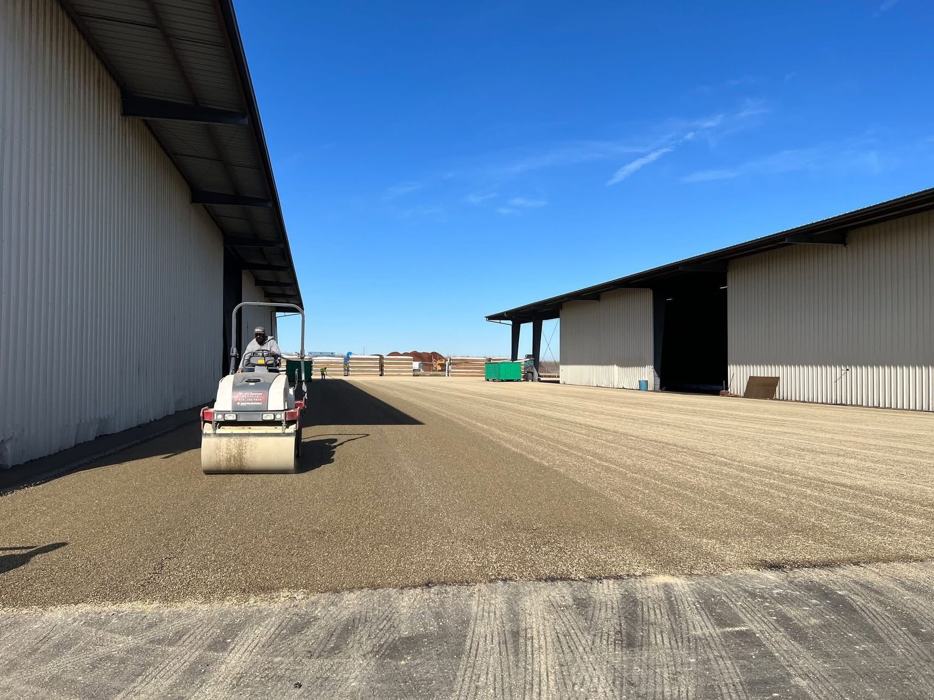 Person driving a small vehicle between large warehouse buildings on a gravel lot under a blue sky