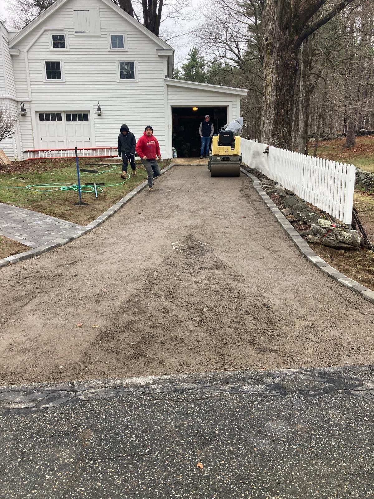 Gravel driveway beside a white house, with two people, a yellow skid steer, and a white fence.