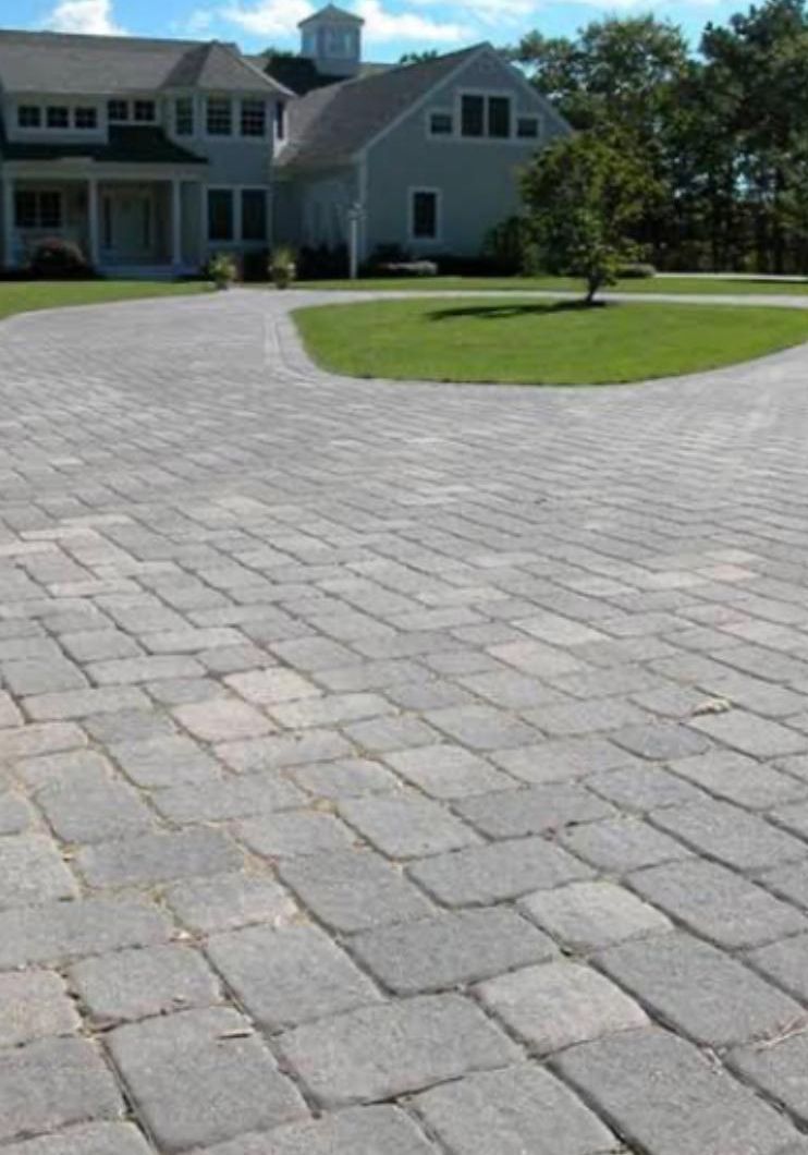 Stone-paver driveway leading to a large gray house with a green lawn and trees.
