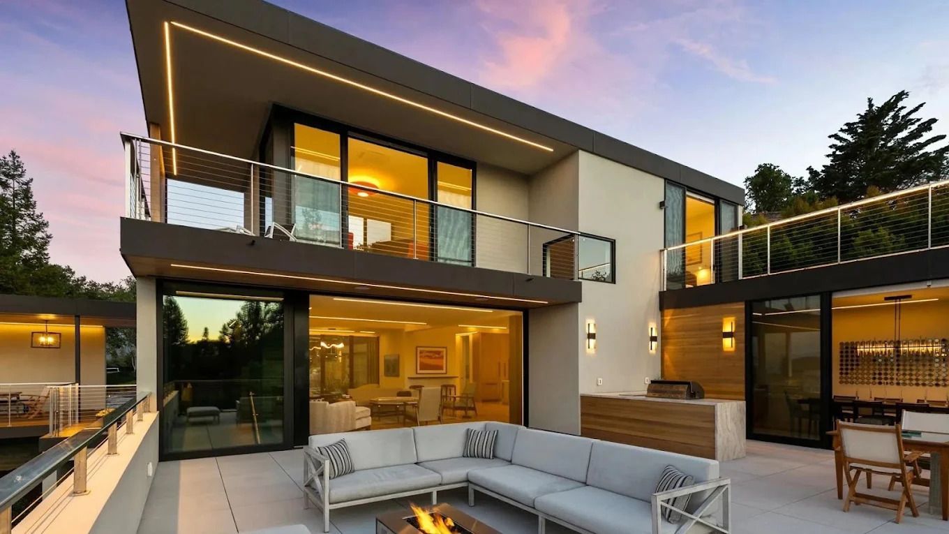 Modern two-story house with glass balconies, warm interior lights, and a patio seating area at dusk