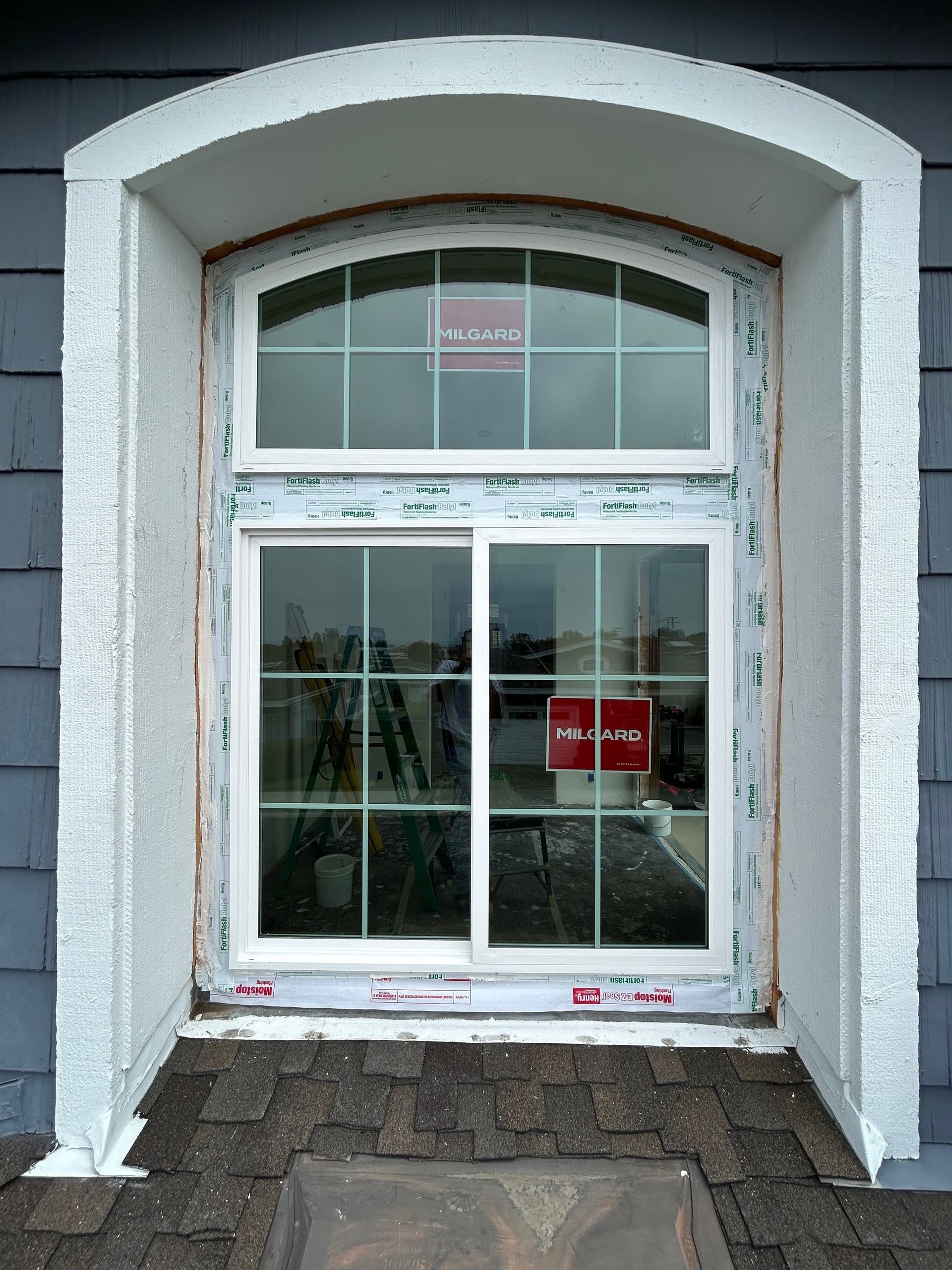 New window installation in an arched alcove with white trim, grids, and protective wrapping against gray siding.