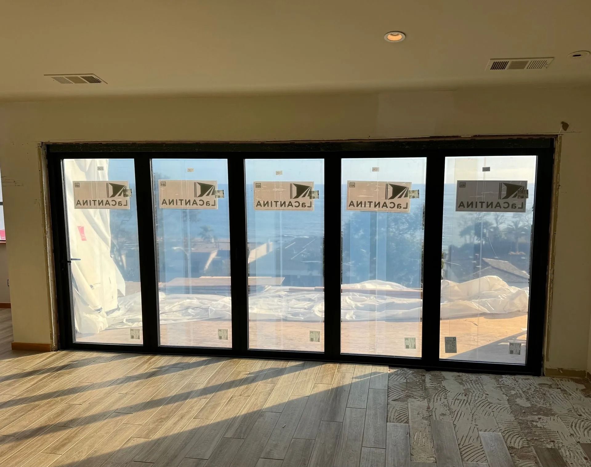 Black framed folding glass doors, uninstalled, in a room with light-colored walls and wood flooring.