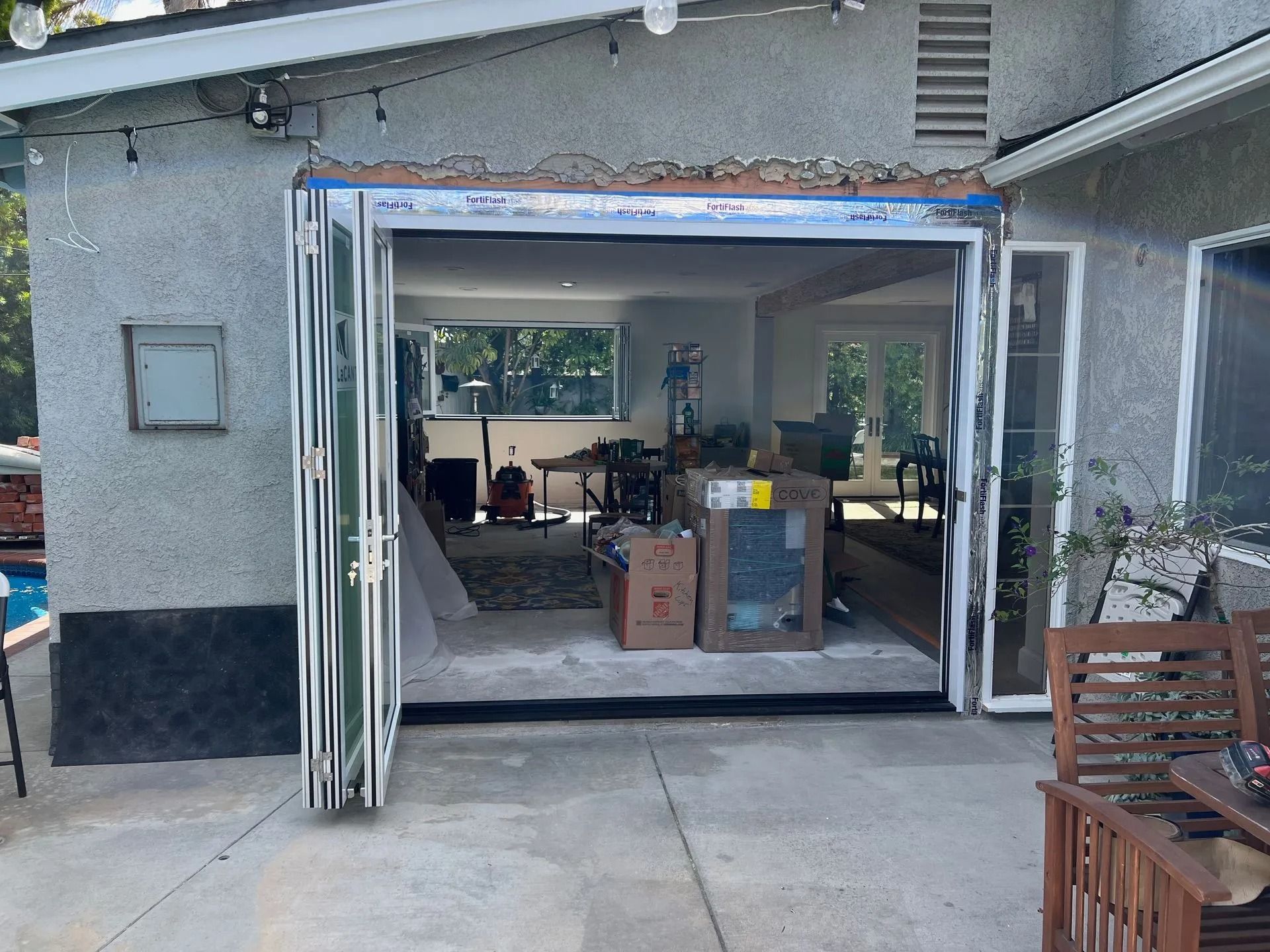 Backyard patio with open folding glass doors revealing interior room with furniture and boxes.