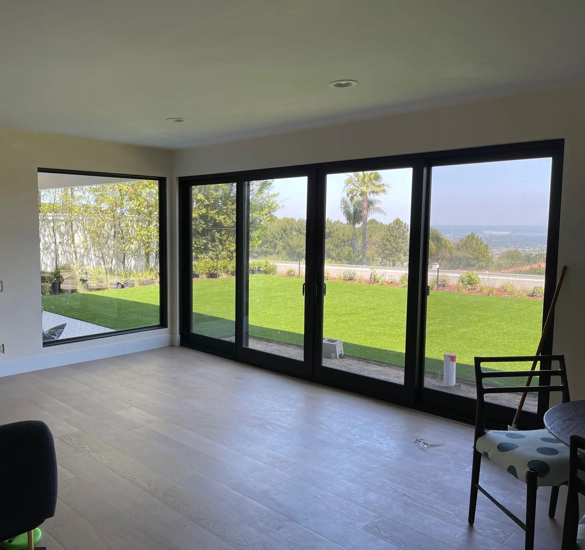 Interior view of room with large windows overlooking a lawn and trees. Black frames, wooden floor.