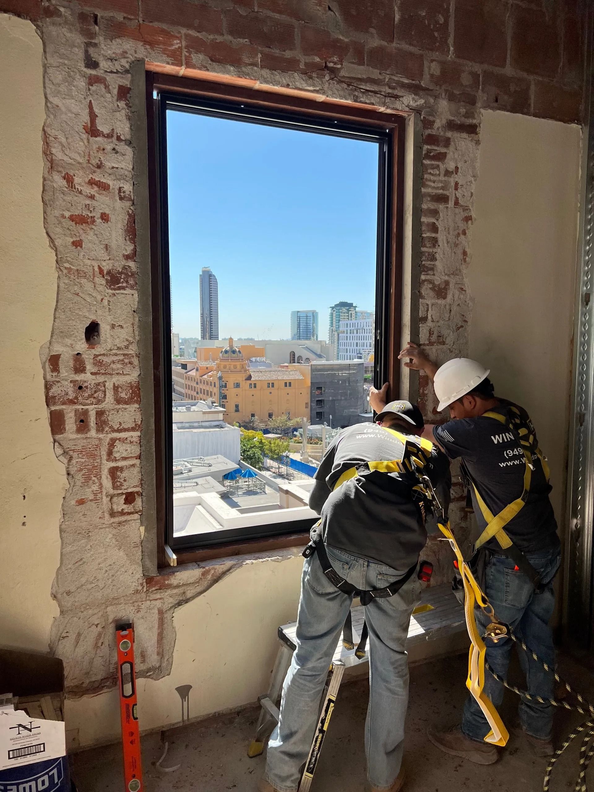 Two construction workers installing a window in a brick-walled building with a city view.