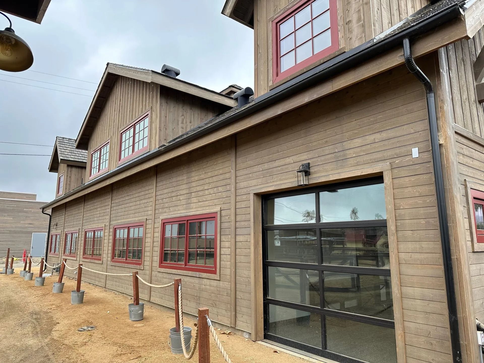 Wooden building with red-framed windows and a glass garage door. A rope fence runs along a dirt path.
