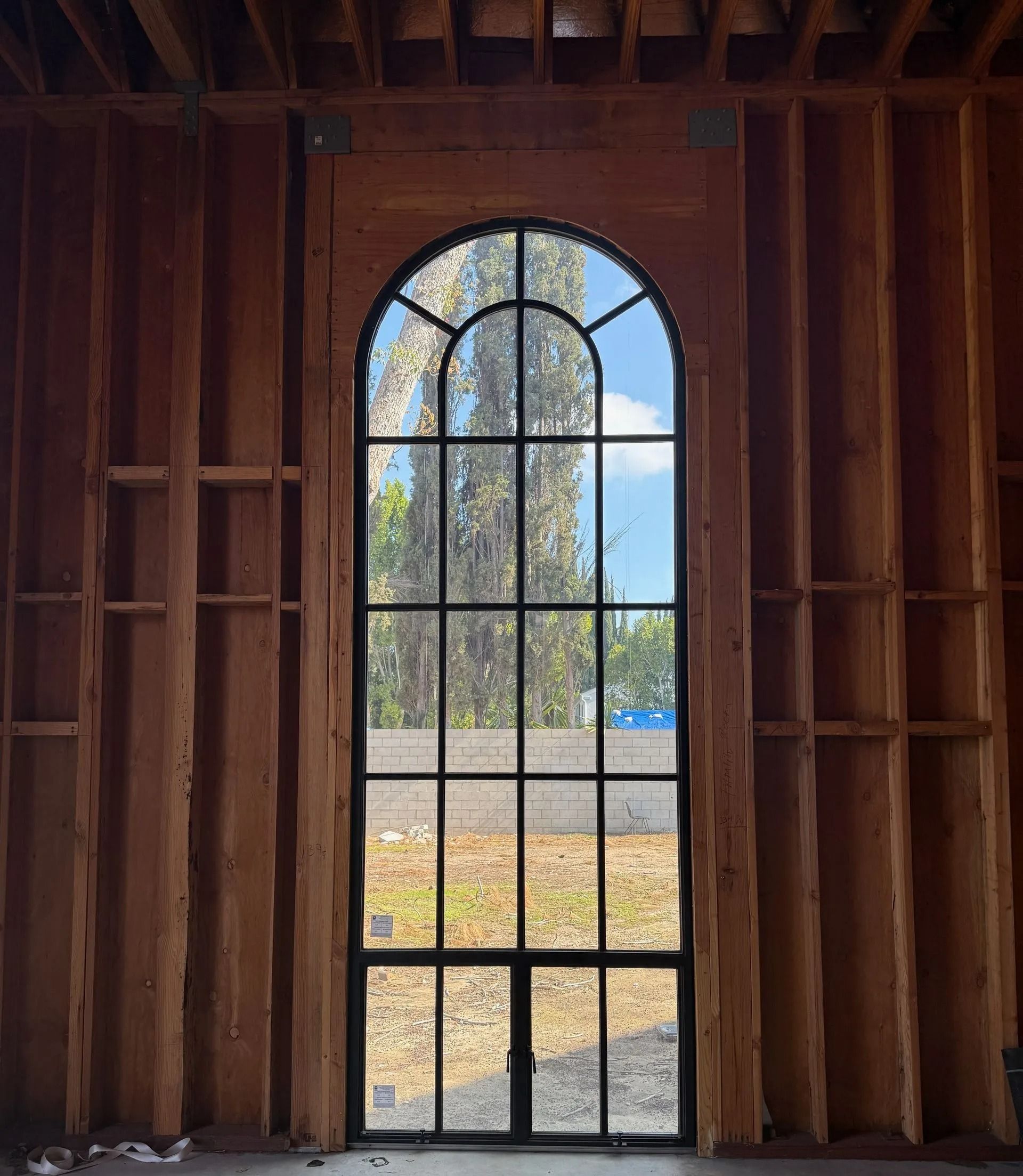 A tall arched window with black gridwork, set in a wood-framed wall, revealing a view of trees and sky.