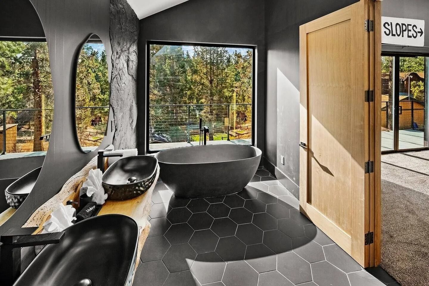 Modern bathroom with black hexagon tiles, gray tub, sinks, and large window overlooking trees.