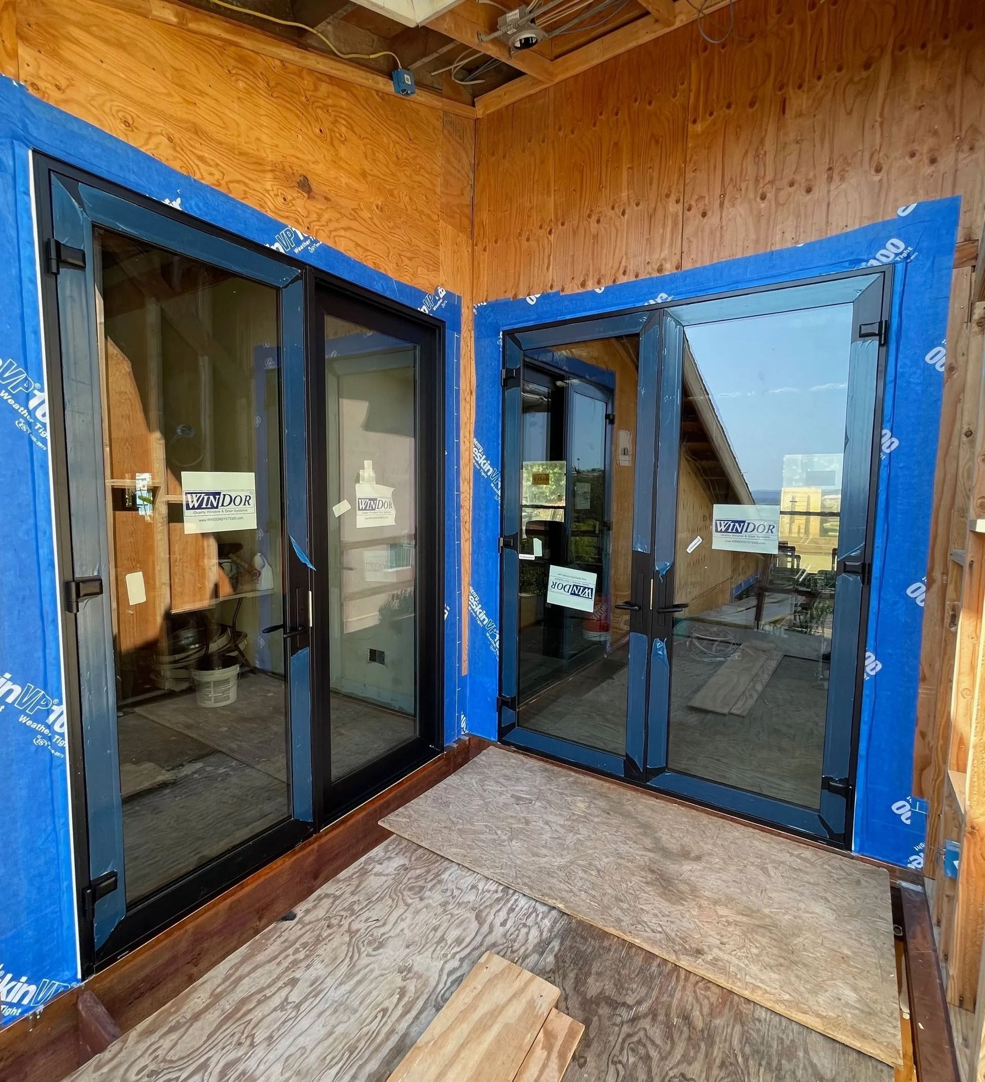 Two glass sliding doors, black frames, blue protective tape. Construction site interior, wood walls, unfinished floor.
