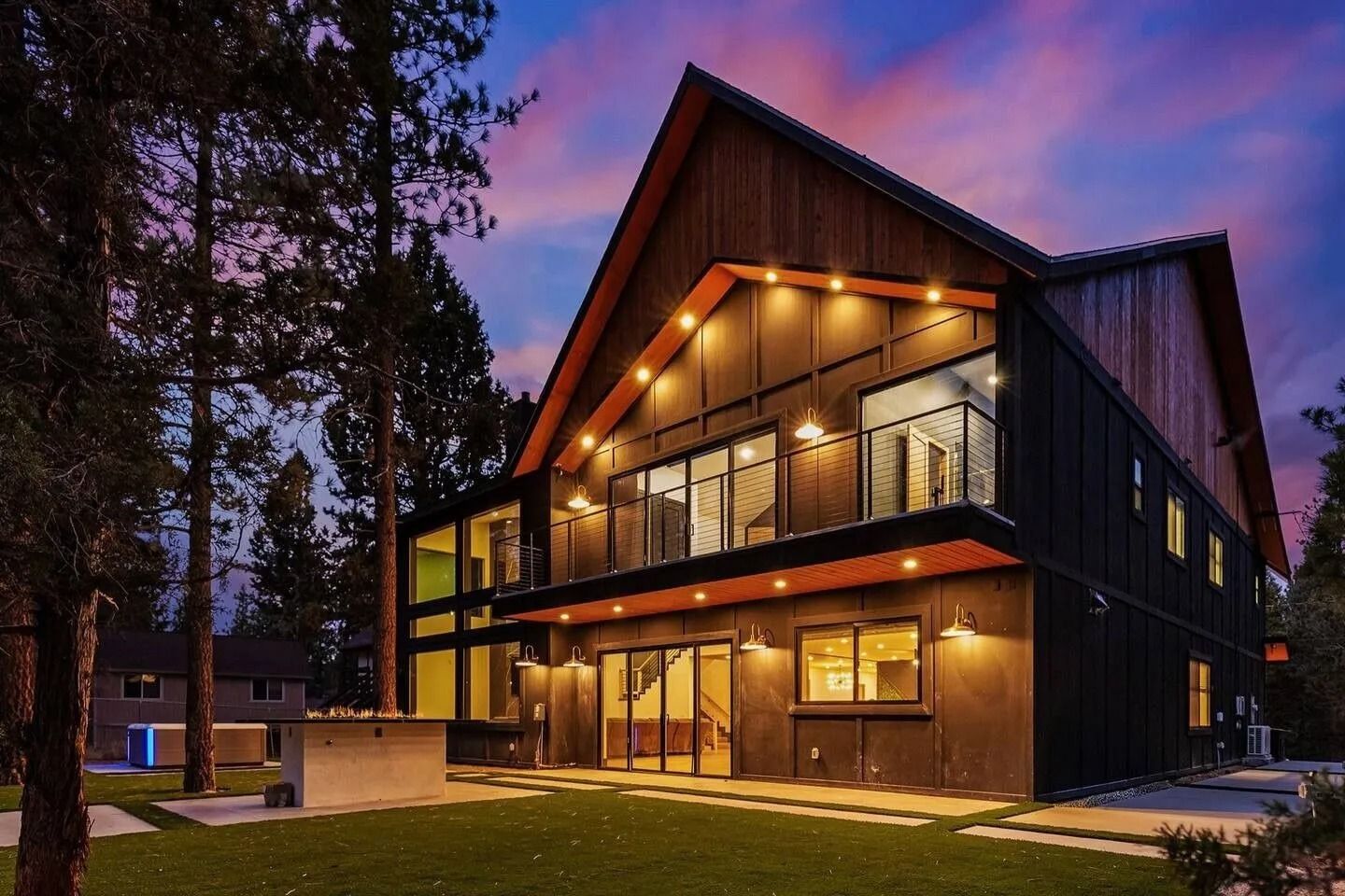 Modern two-story house with dark siding and large windows, lit up at dusk with a purple and blue sky.