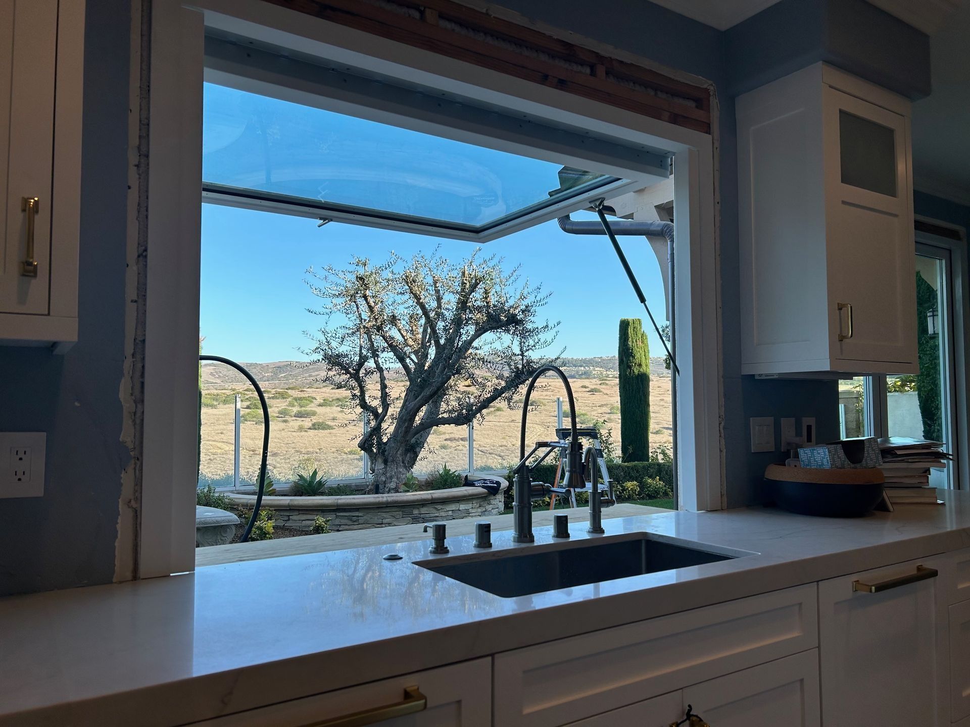 Kitchen sink with an open window, overlooking a tree in a field under a blue sky.