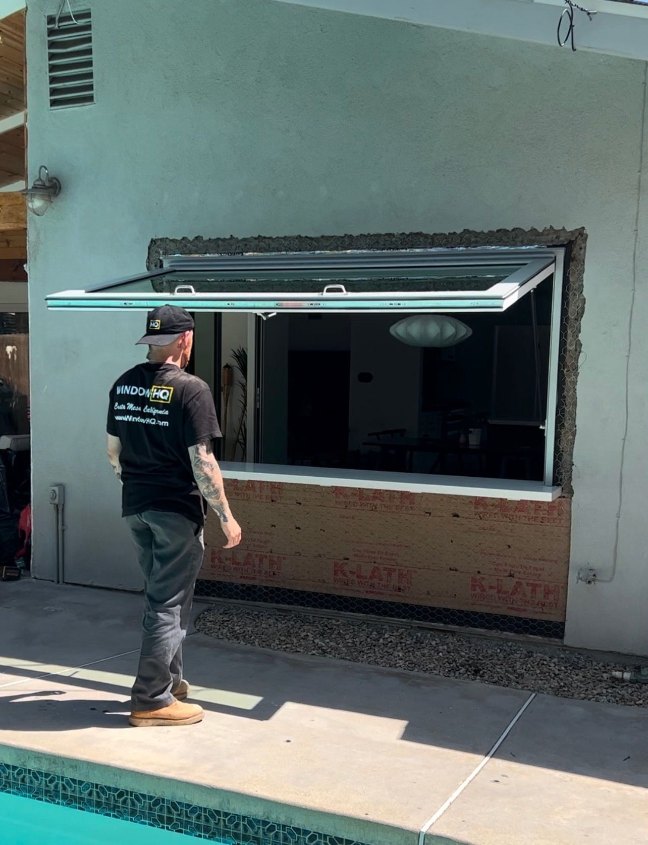 Man opens up an outdoor window to reveal an interior space. The window is above a brick ledge, next to a pool.