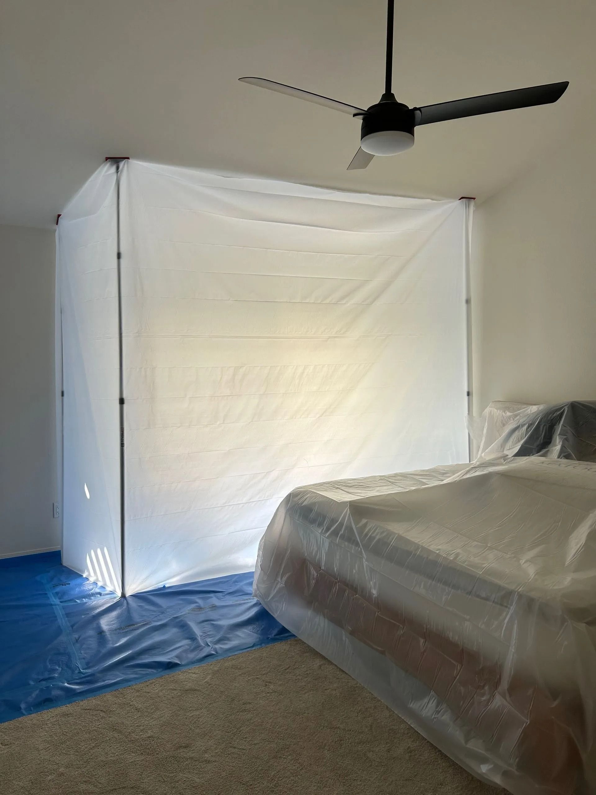 Room with a plastic-covered bed and a containment structure made of poles and white plastic sheeting.