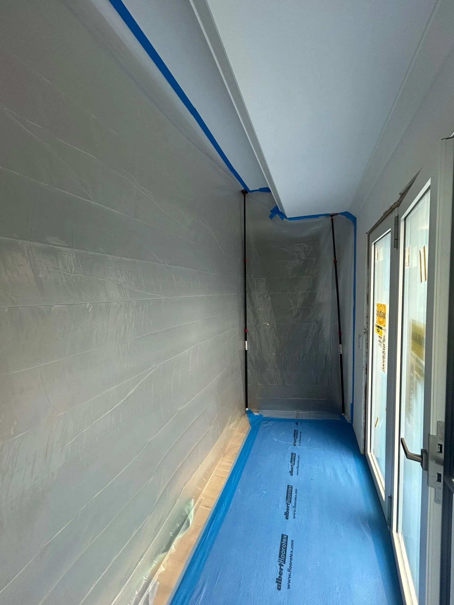 Narrow room with protective blue tape and flooring. White walls, doors on the right.