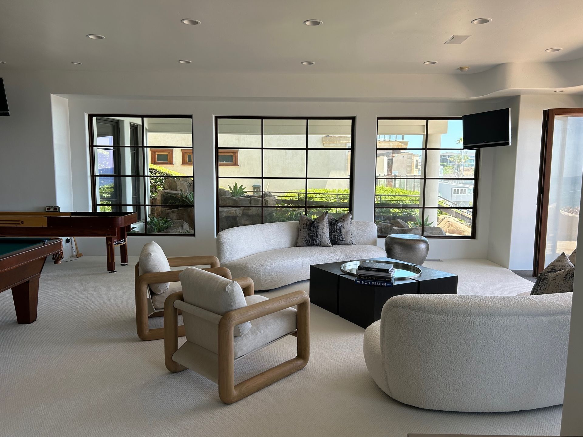Living room with white walls, black-framed windows, white couches and chairs, black coffee table, and a pool table.