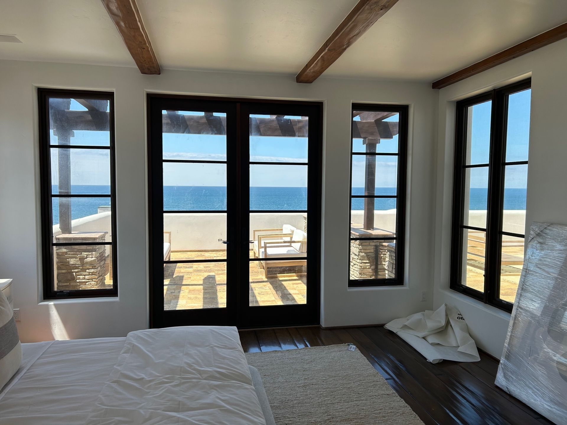 Bedroom with ocean view; black-framed windows and doors, white walls, wooden beams, rug, and sunlight.