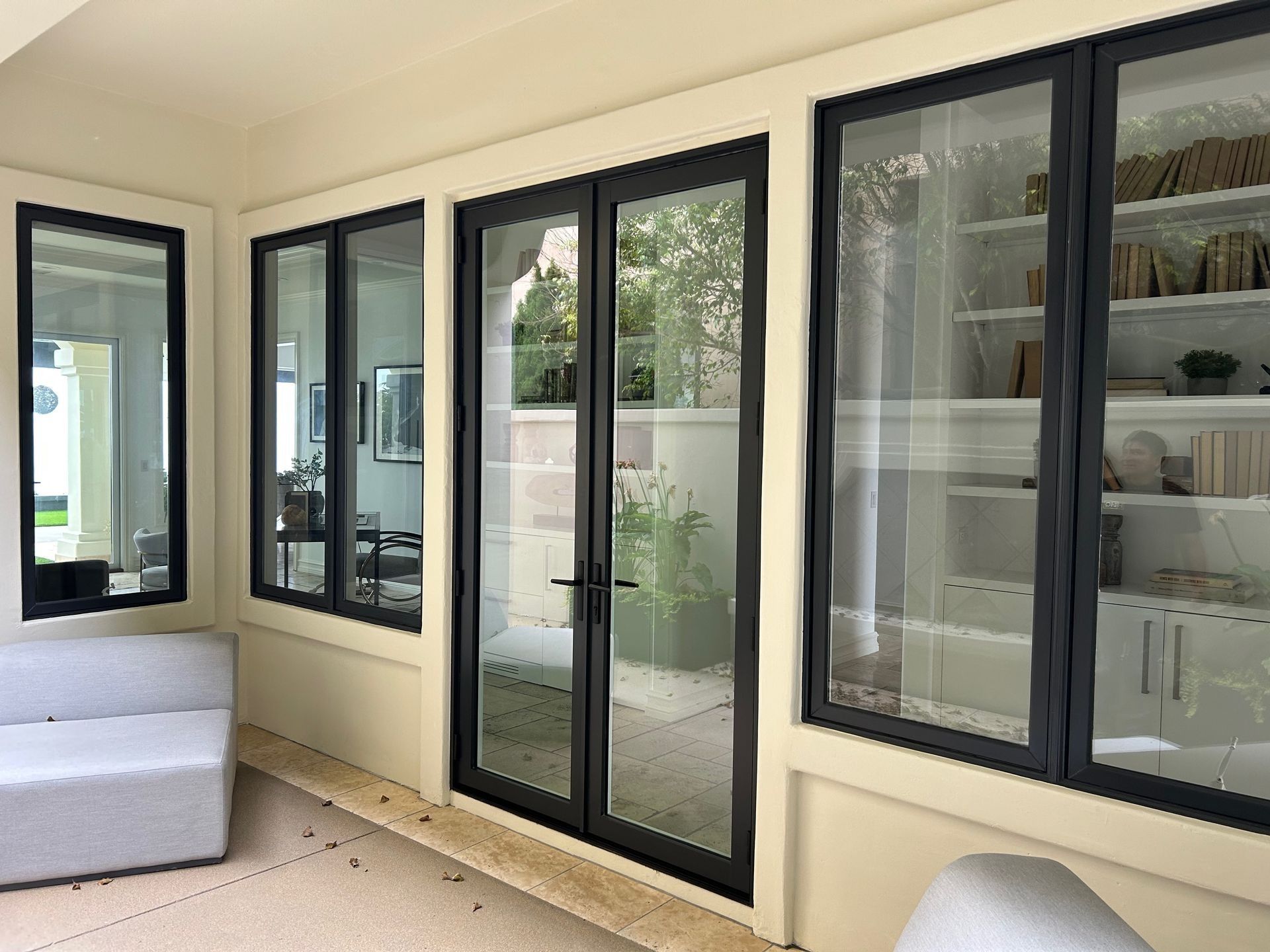 Sunroom with black-framed windows and doors, looking into a garden and a room with shelves. Beige walls and floor.