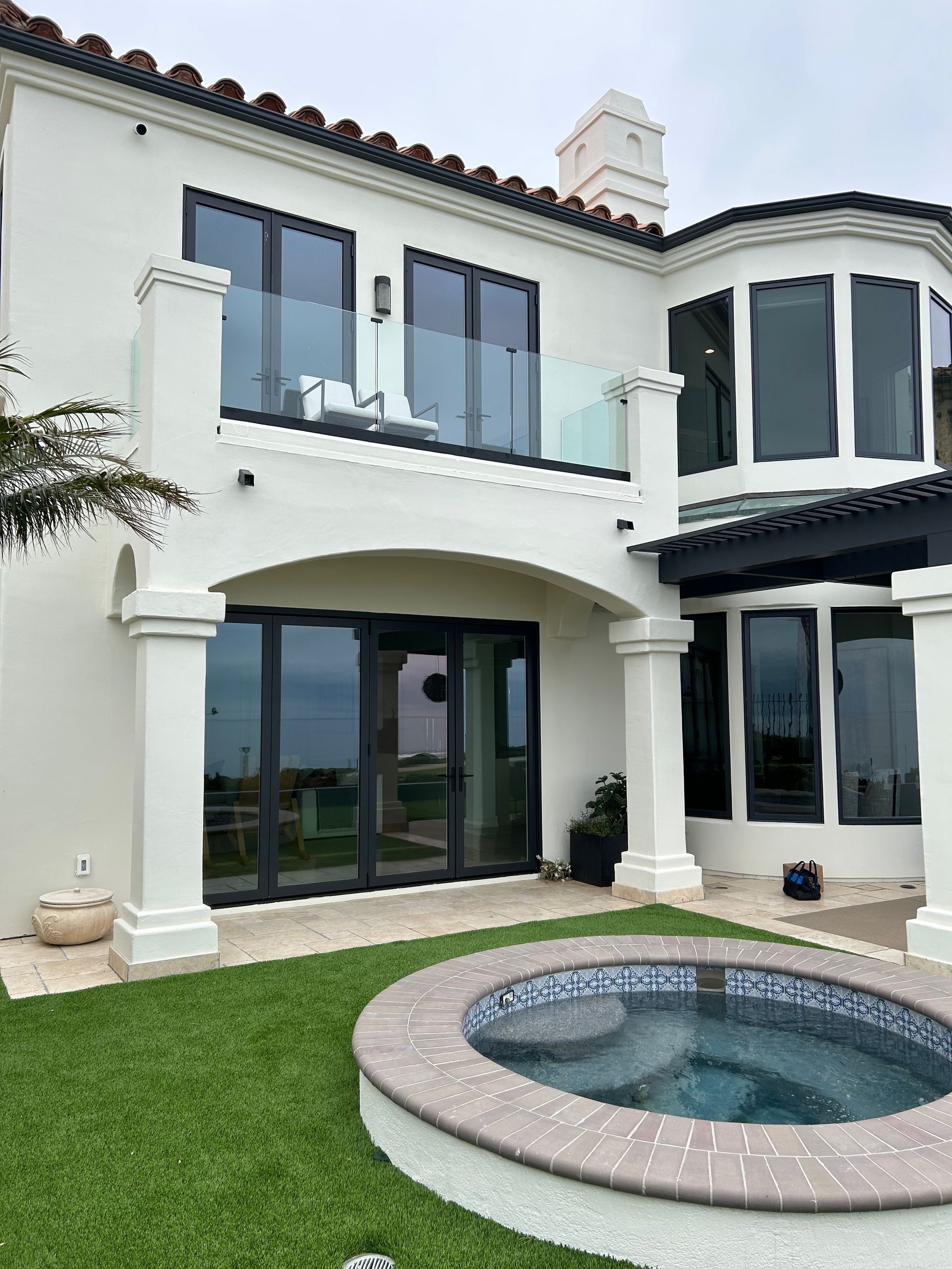 White house with black-framed windows and a small spa. Balcony with glass railing, green grass lawn.