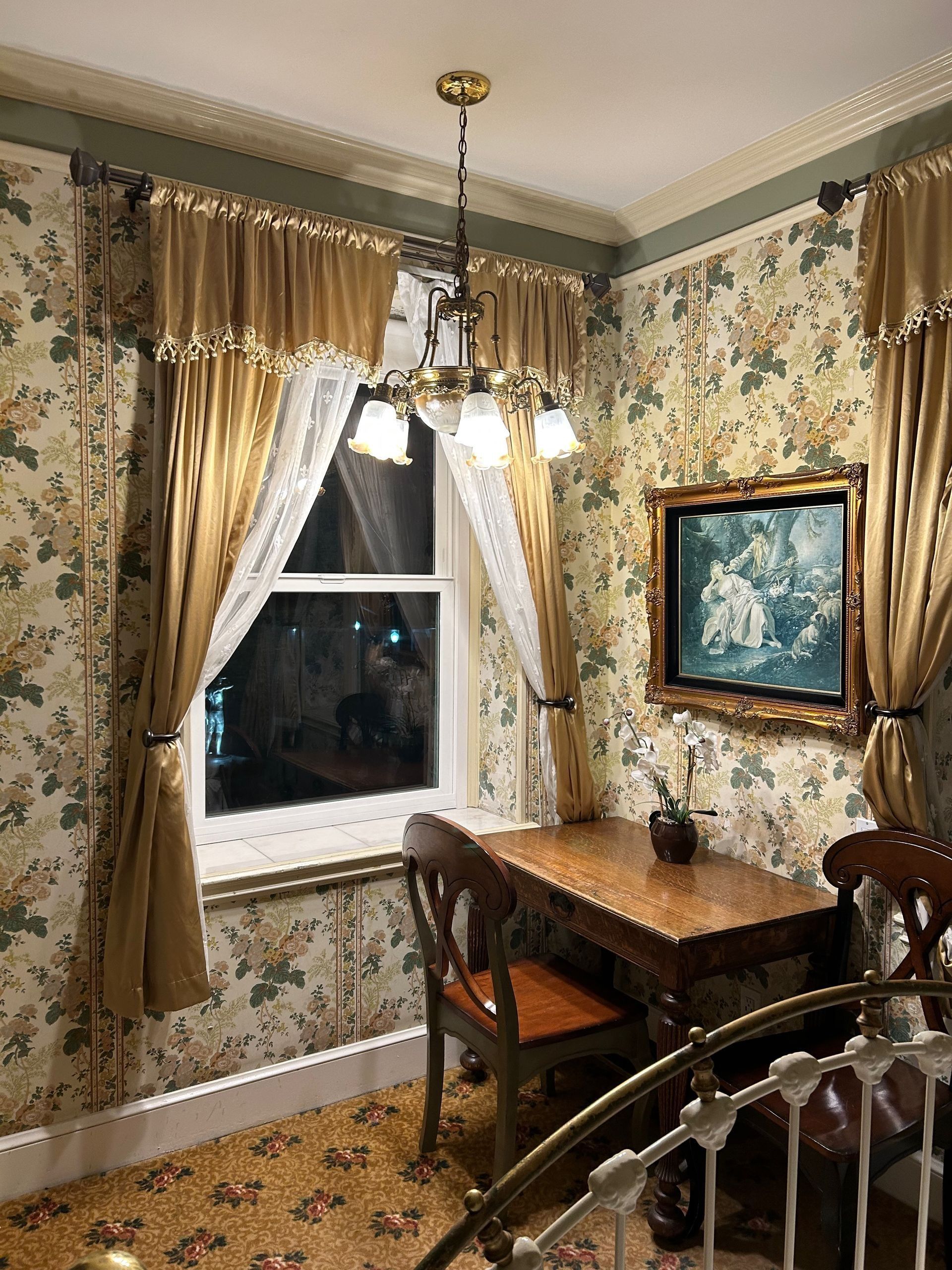 A room with a desk, window, curtains, floral wallpaper, and a chandelier.