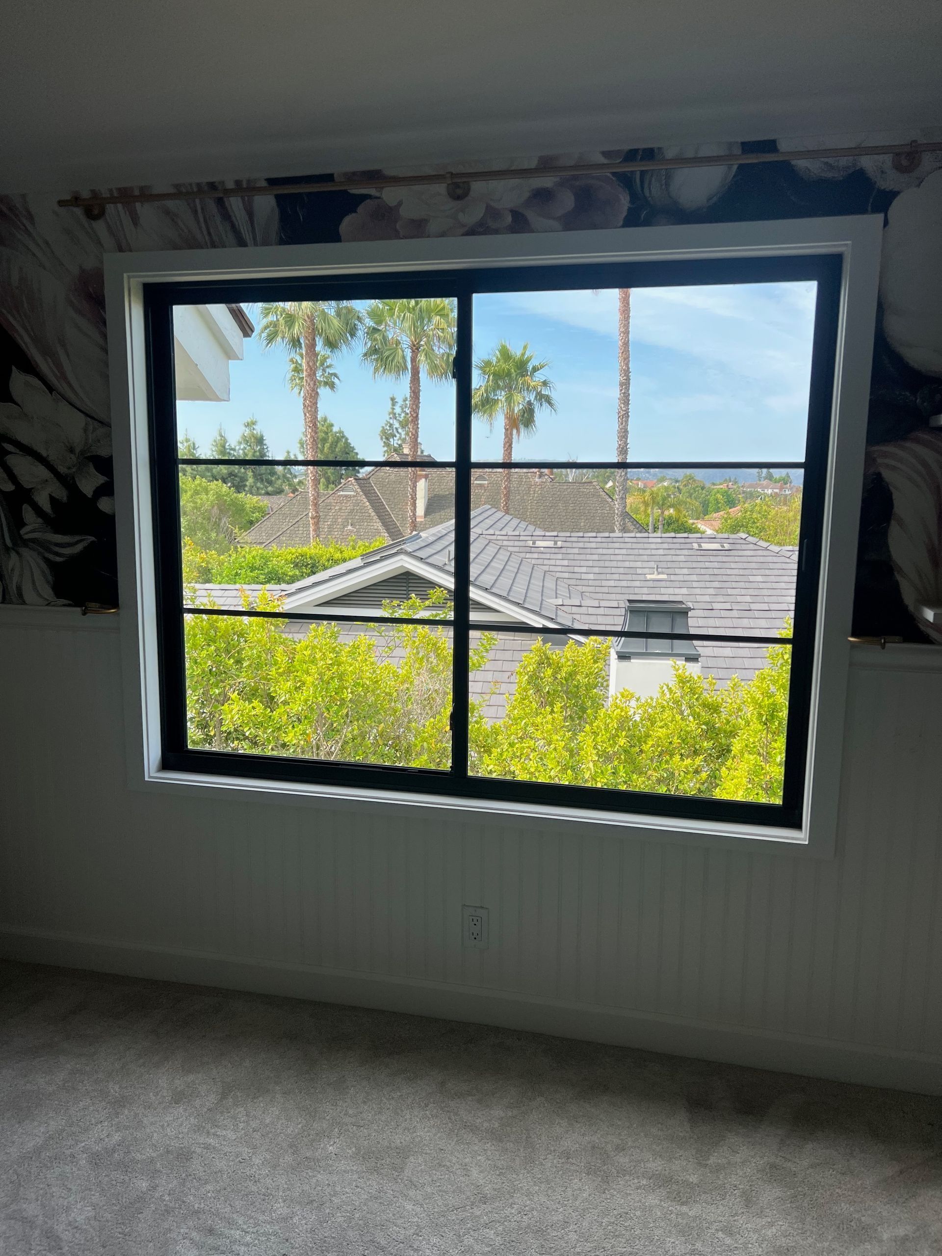 Black-framed window with view of rooftops, palm trees, and blue sky. Interior room with white walls, carpet, and faded wallpaper.