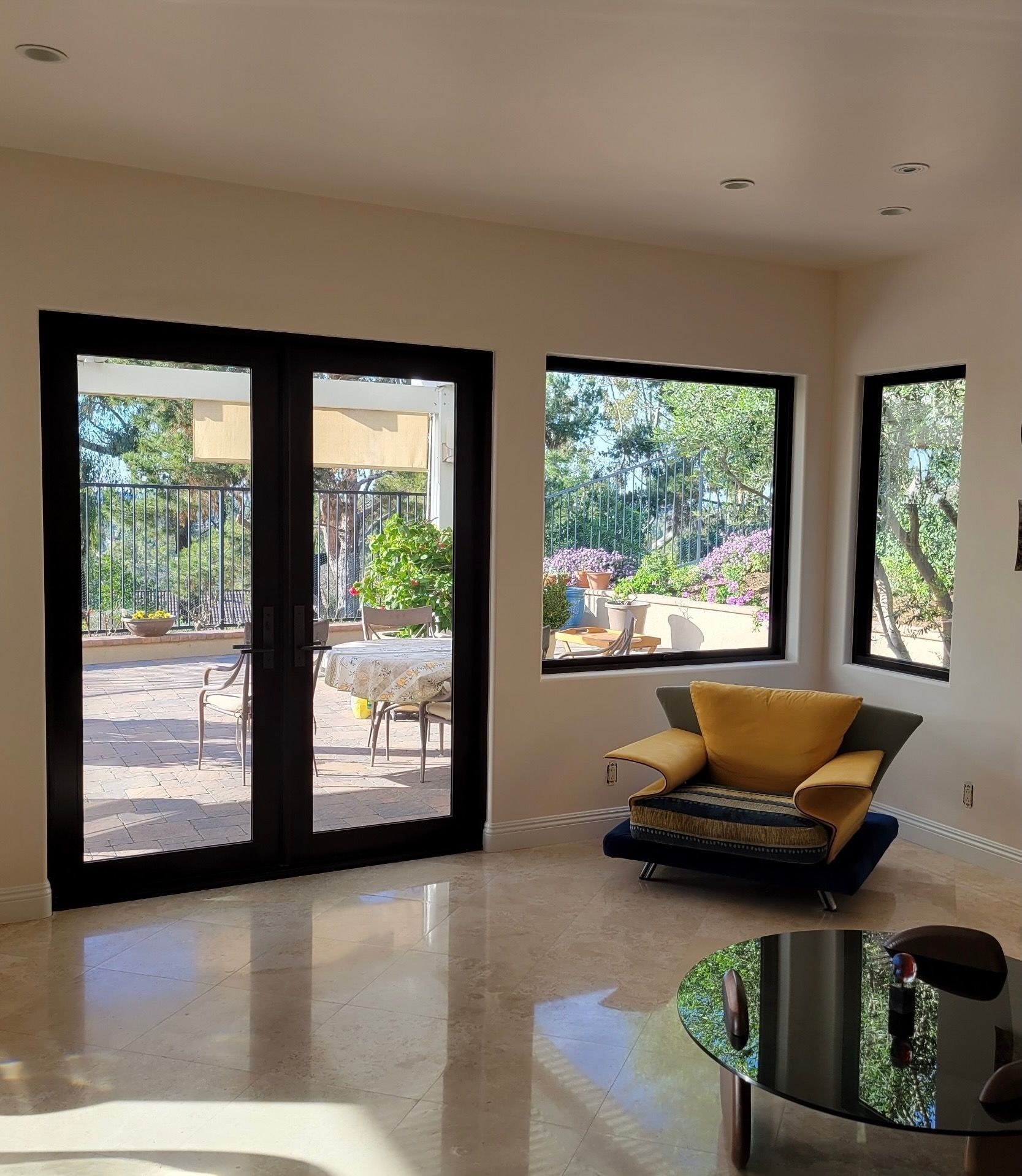 Living room with French doors and two windows; overlooks a patio and garden. A blue and yellow chair and a glass coffee table are in the room.
