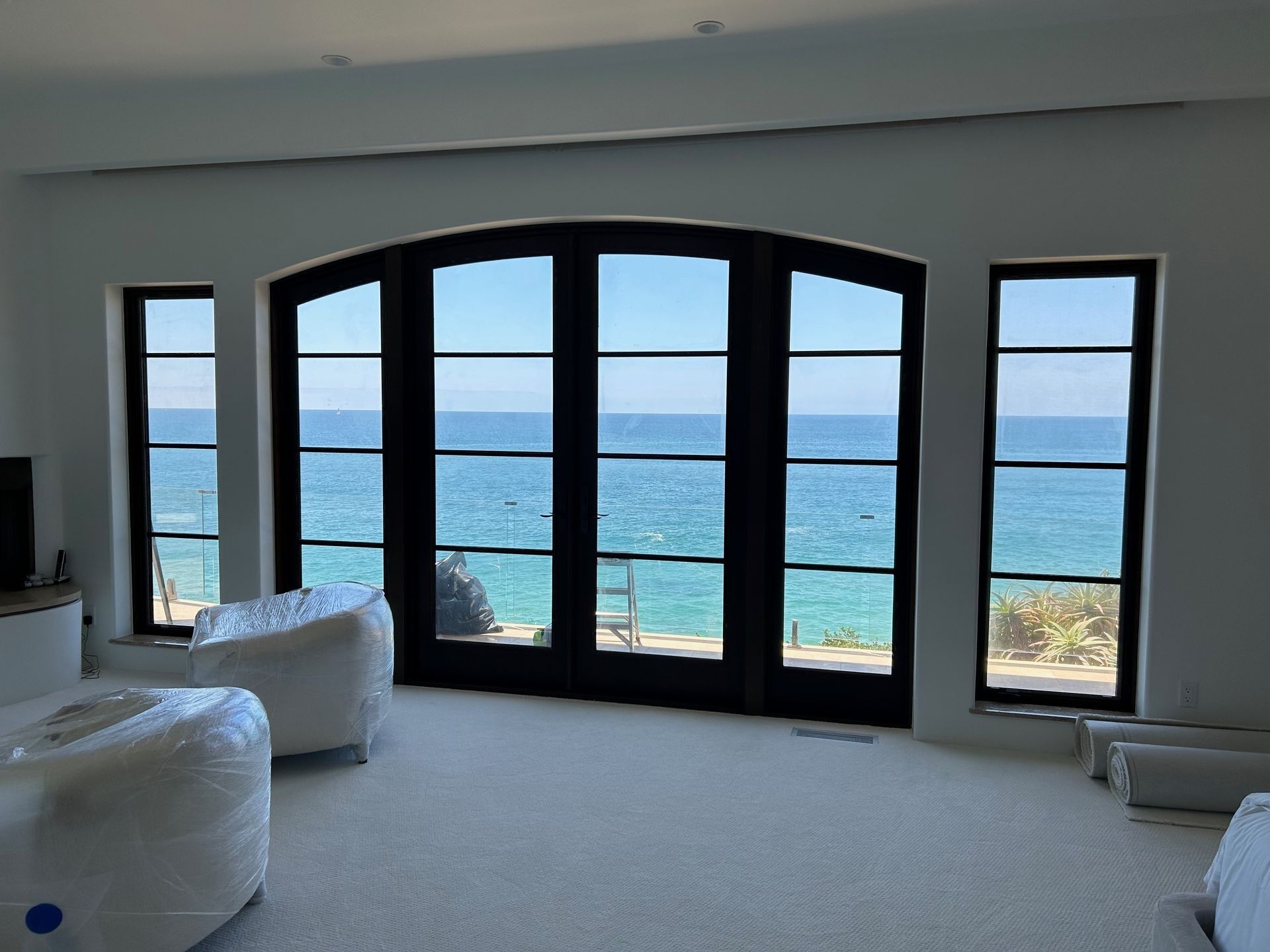 Room with large black-framed windows overlooking the ocean. Two white-wrapped chairs in the foreground.