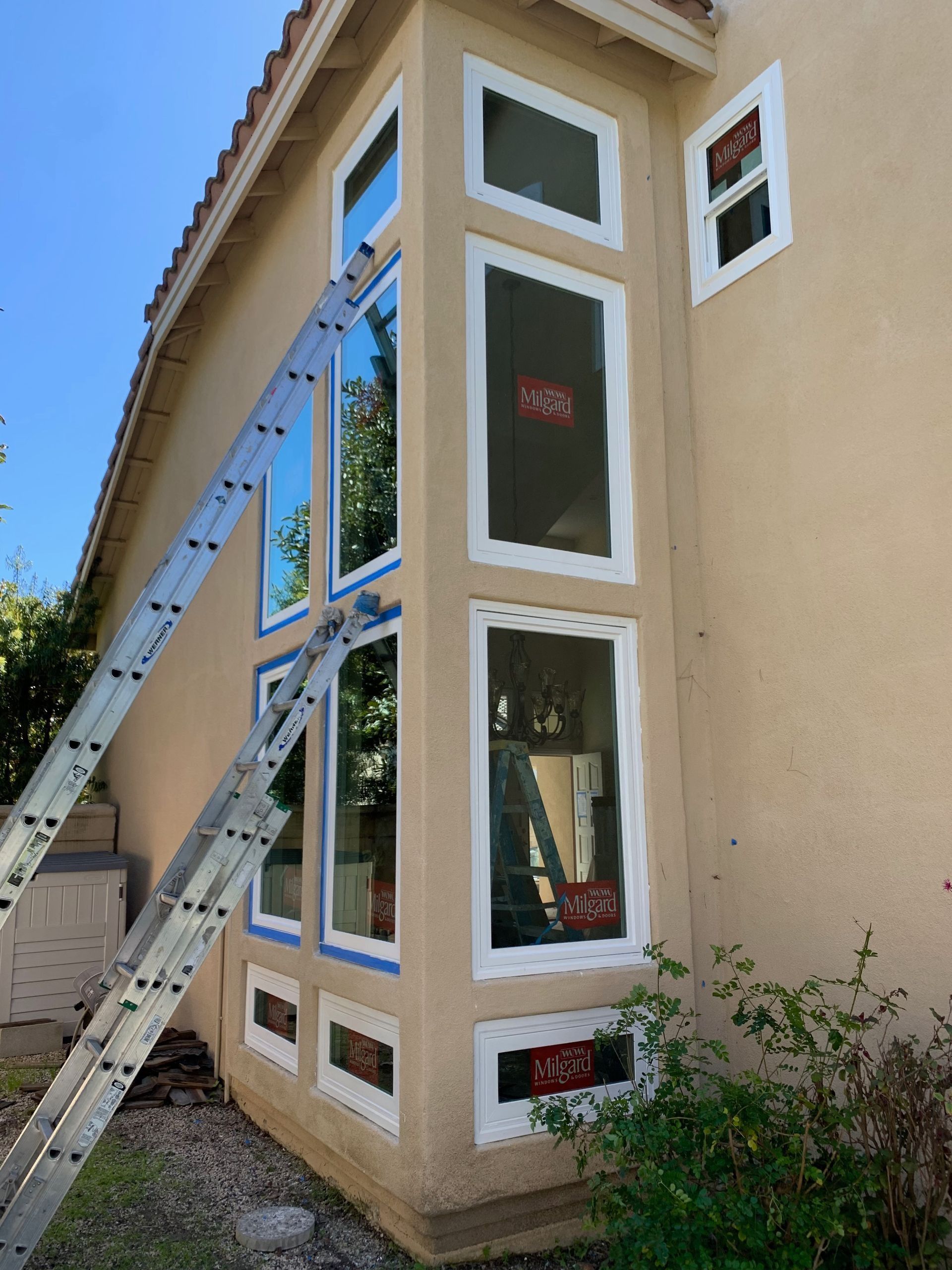 Ladder leaning against a beige house with tall, white-framed windows; some windows have stickers.