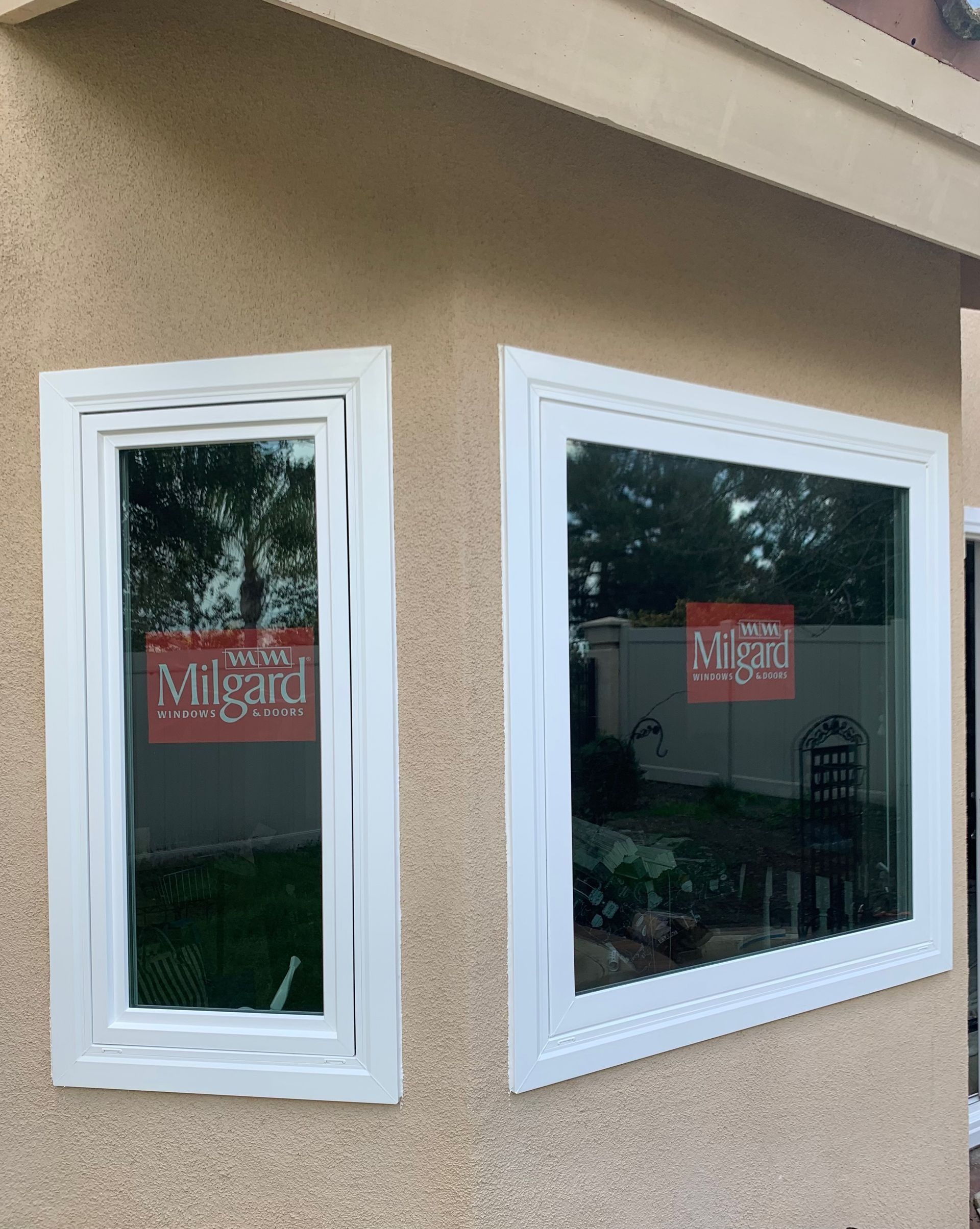 Two white-framed Milgard windows on a beige stucco wall, reflecting trees and grass.